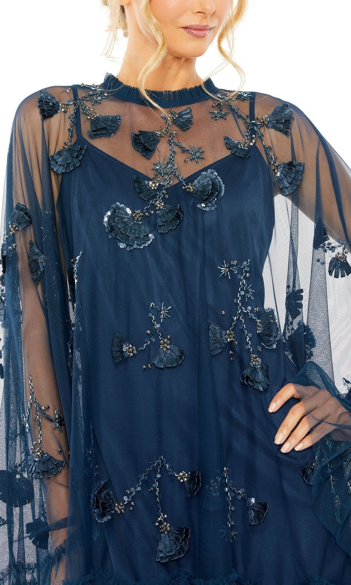 Woman in a Mac Duggal 9257 Long Sleeve Short Hoco Dress, navy blue with sheer fabric and floral leaf appliqués, poses stylishly with one hand on her hip.