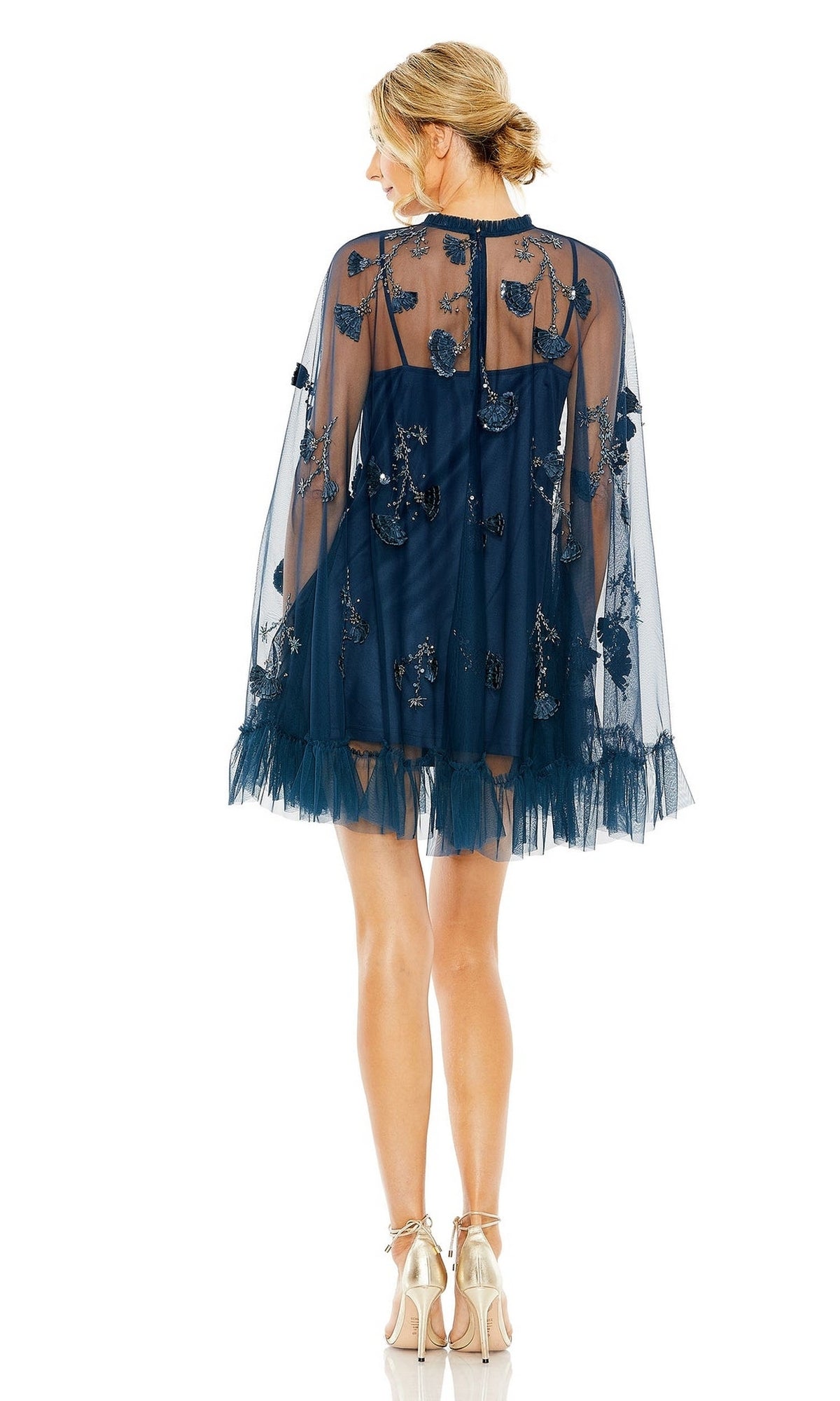 Woman in a navy blue sheer Long Sleeve Short Hoco Dress: Mac Duggal 9257 with floral embroidery and ruffle hem, paired with beige heels, stands facing away from the camera against a white background.