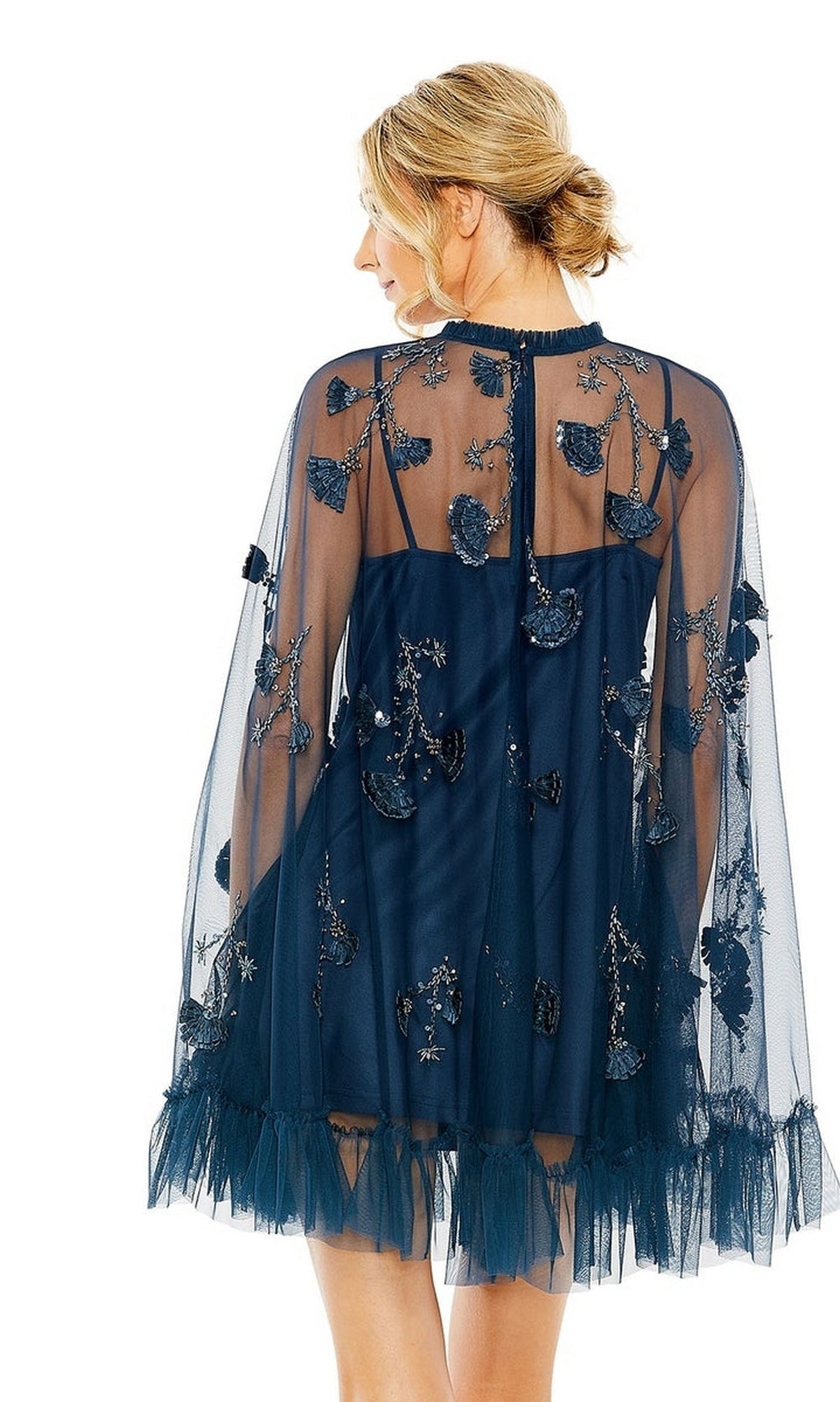 A woman is seen from the back wearing the Mac Duggal 9257 Long Sleeve Short Hoco Dress in dark blue with sheer fabric, floral embroidery, and tulle details, set against a white background.