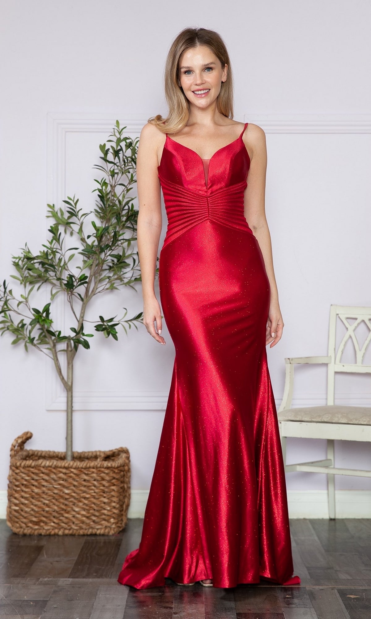 A woman smiles at the camera indoors, standing near a potted plant and white chair, wearing the Sheer Cut-Out Sides Long Prom Dress 9260—a shiny red v-neck evening gown.