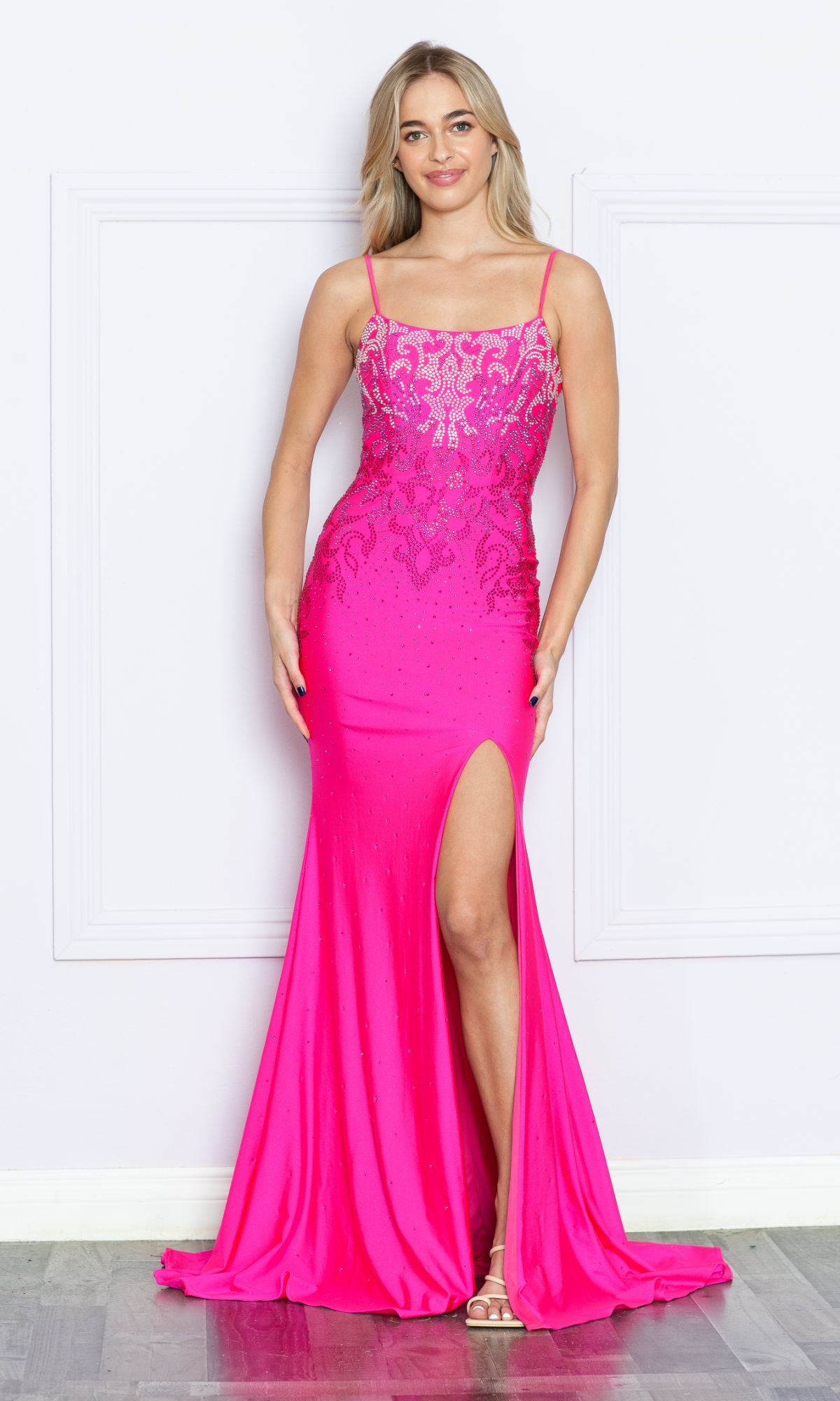 A woman stands against a white wall wearing the Heat-Stone Ombre Long Prom Dress 9264—a bright pink, floor-length gown with thin straps, intricate lace details, and a high slit.