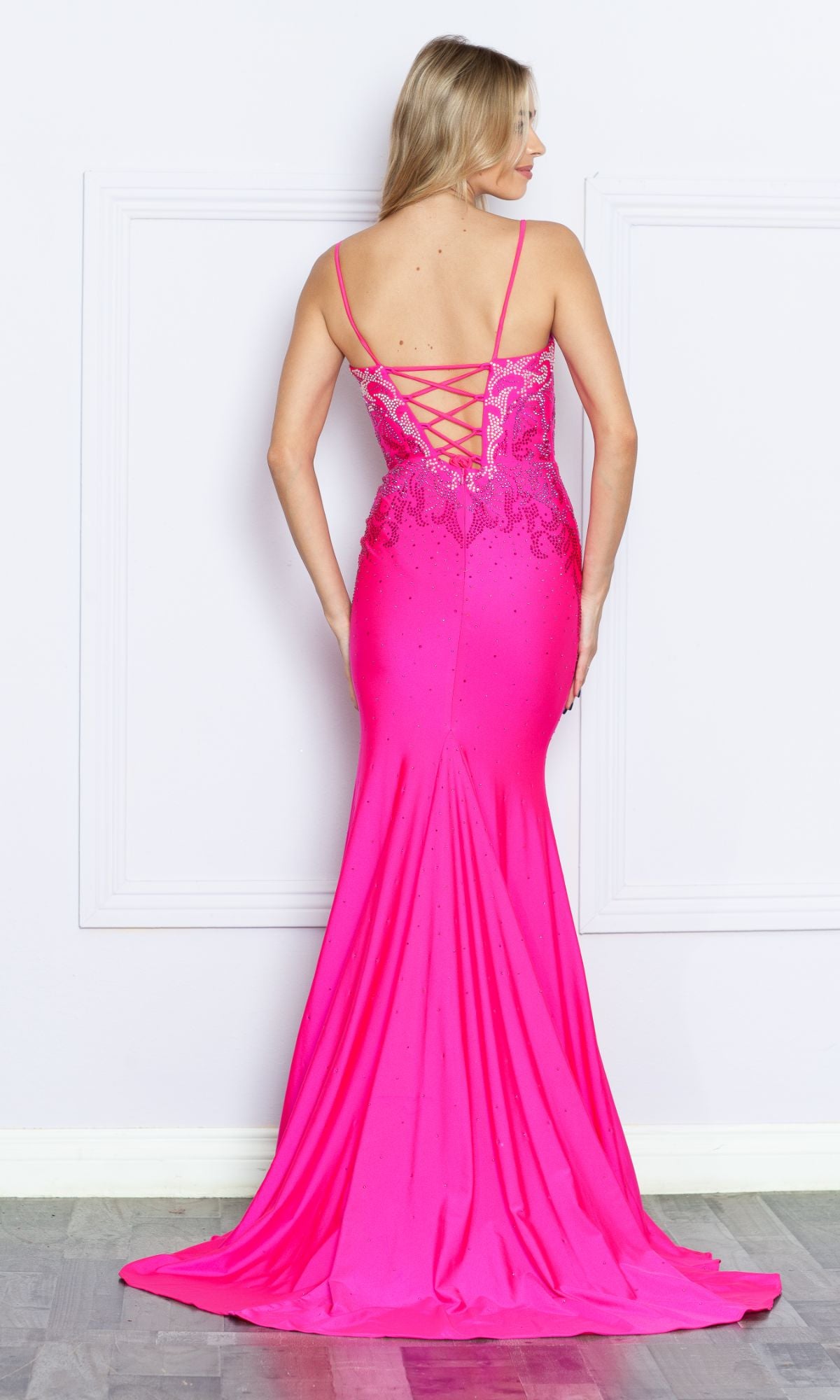 A woman stands facing a white wall, wearing the Heat-Stone Ombre Long Prom Dress 9264—a bright pink, floor-length gown with thin straps and a lace-up back.