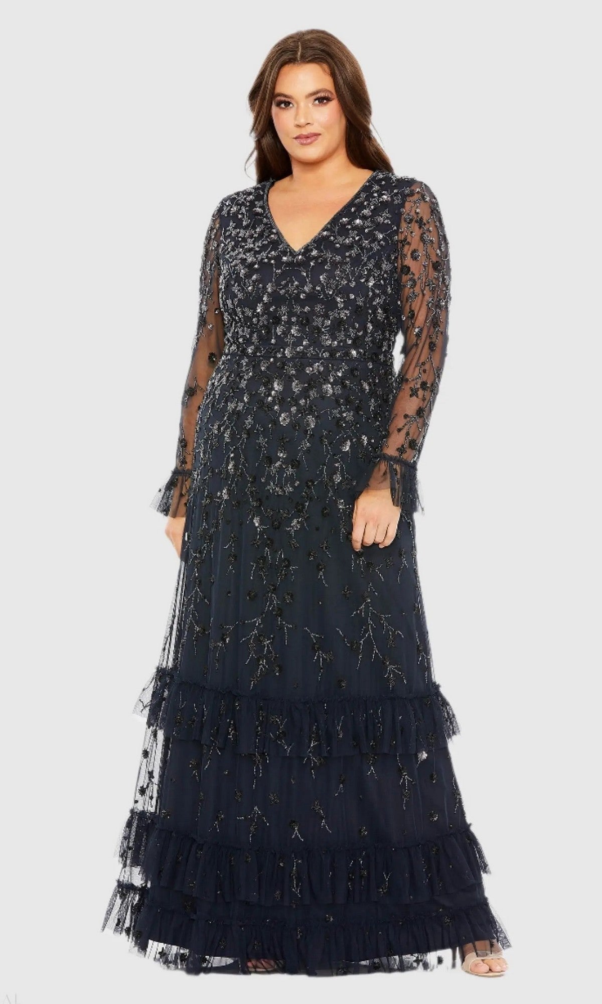 A woman wearing the Mac Duggal 9271 long plus-size formal dress in Midnight, featuring floral embroidery, sheer long sleeves, a V-neckline, and a tiered ruffle skirt—a stunning choice for anyone seeking an elegant plus-size evening gown.