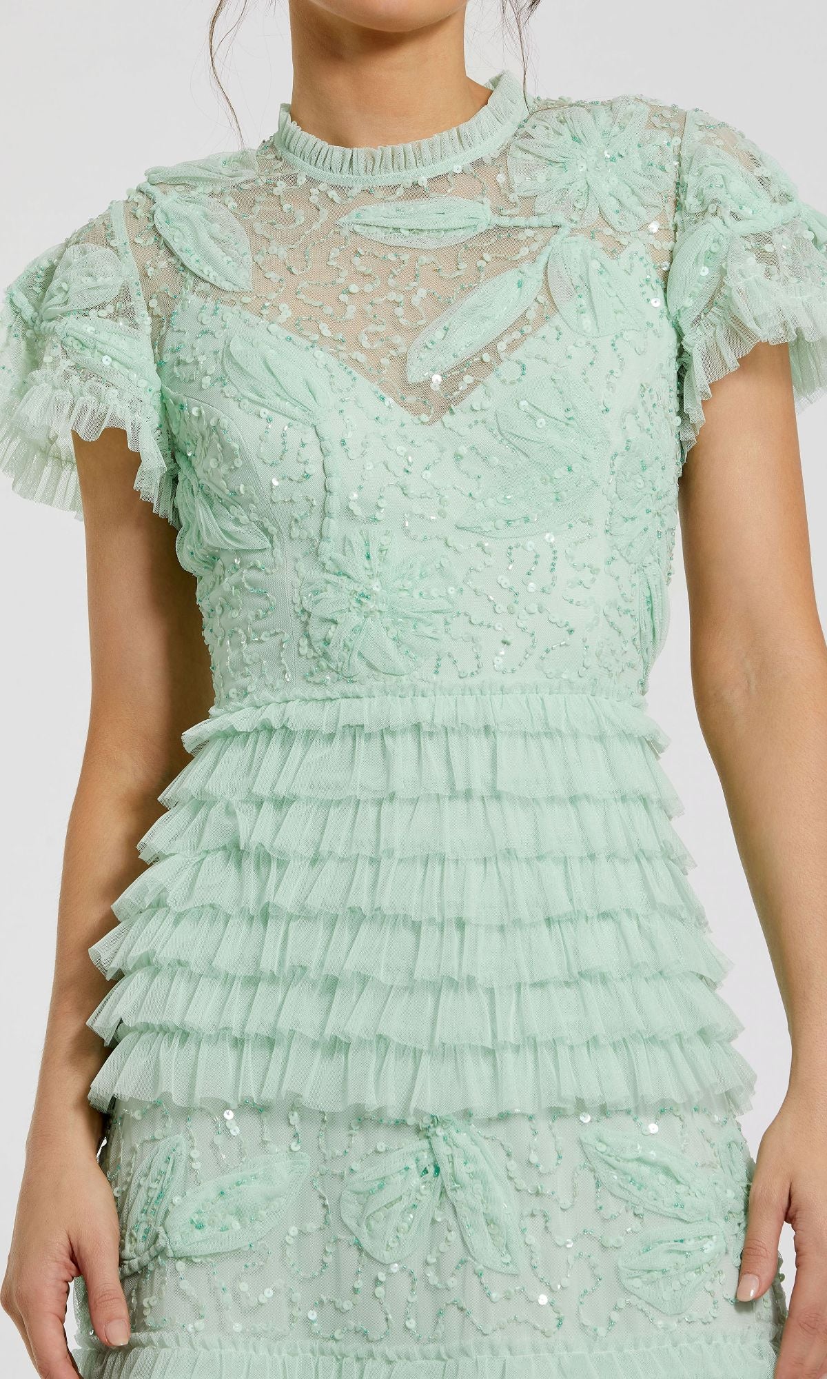 Woman in a light sage green evening gown featuring sheer fabric, floral appliqué, sequins, and layered ruffles on the sleeves and waist—a look like the Mac Duggal 9278 Long Sage Green Formal Dress.