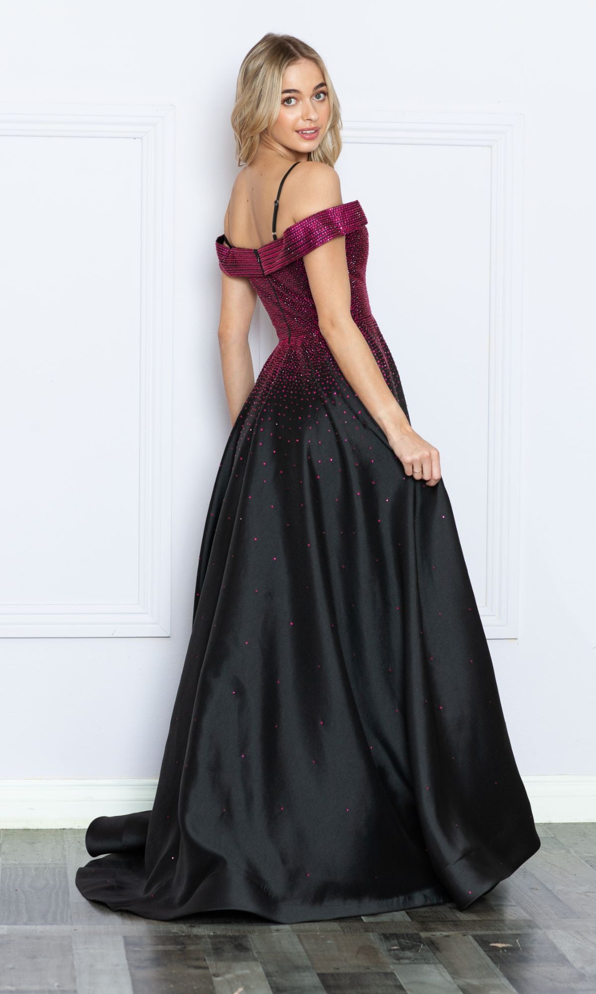 A woman in the Cold-Shoulder Beaded Long Prom Ball Gown 9278, featuring a magenta fitted bodice and floor-length skirt, stands facing away and glances over her shoulder.