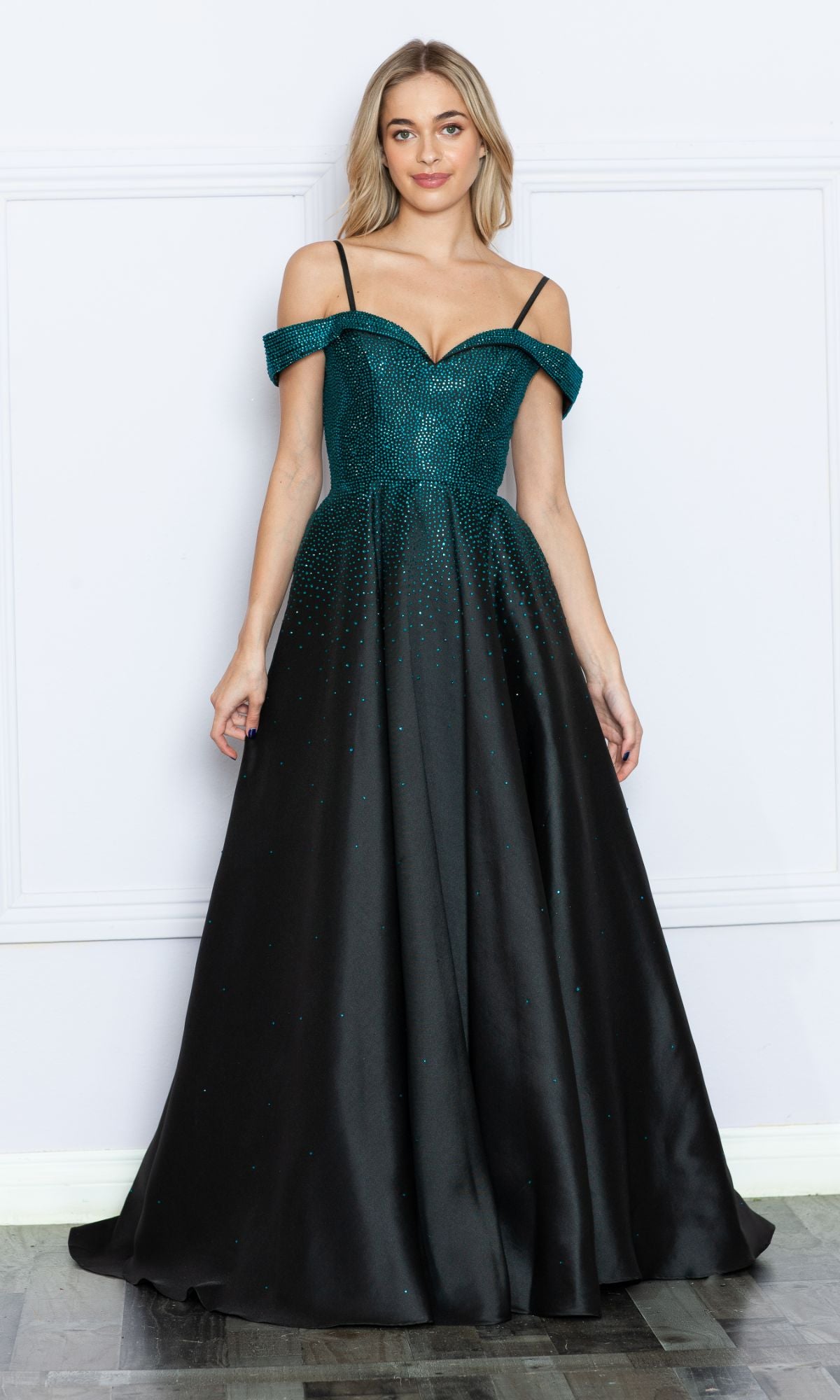 A woman stands against a white wall wearing the Cold-Shoulder Beaded Long Prom Ball Gown 9278, a floor-length dark green and black gown with thin straps, off-the-shoulder design, sparkling beads, and a fitted bodice.