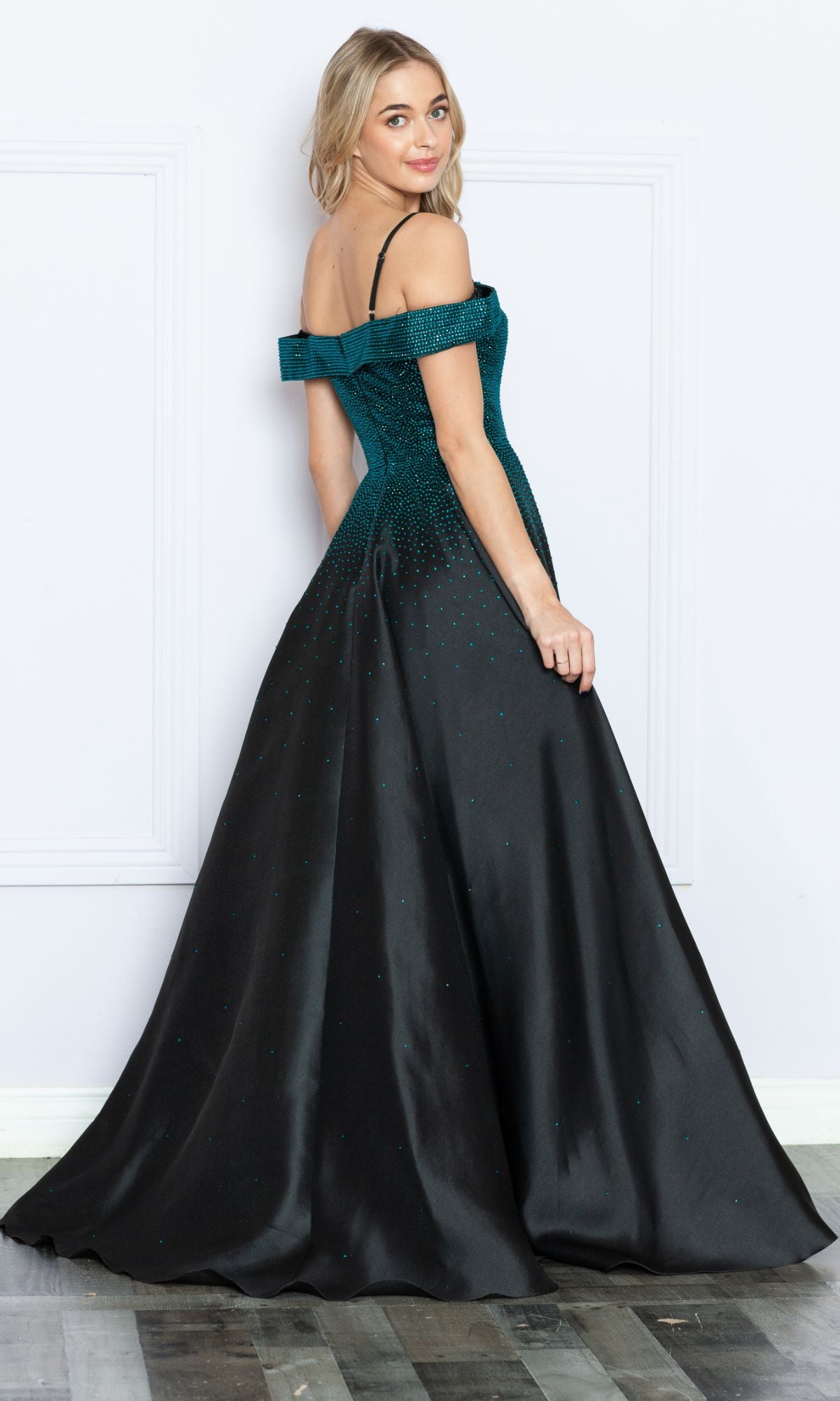 Woman wearing the Cold-Shoulder Beaded Long Prom Ball Gown 9278, a long A-line teal and black dress with flared skirt, stands on a wood floor against a white background, looking over her shoulder.