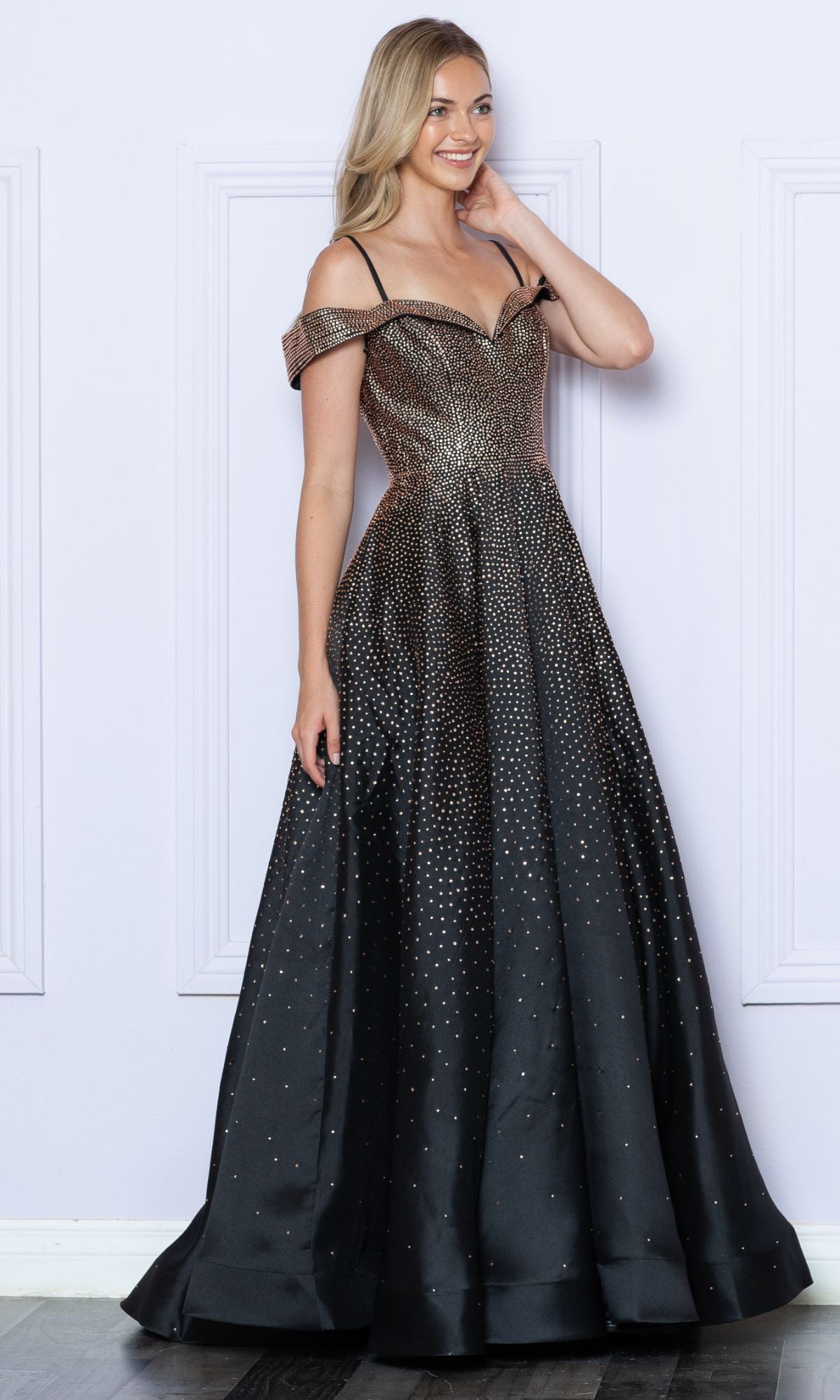 A woman in the Cold-Shoulder Beaded Long Prom Ball Gown 9278, featuring sparkling black beads and thin straps, smiles while standing against a white wall.