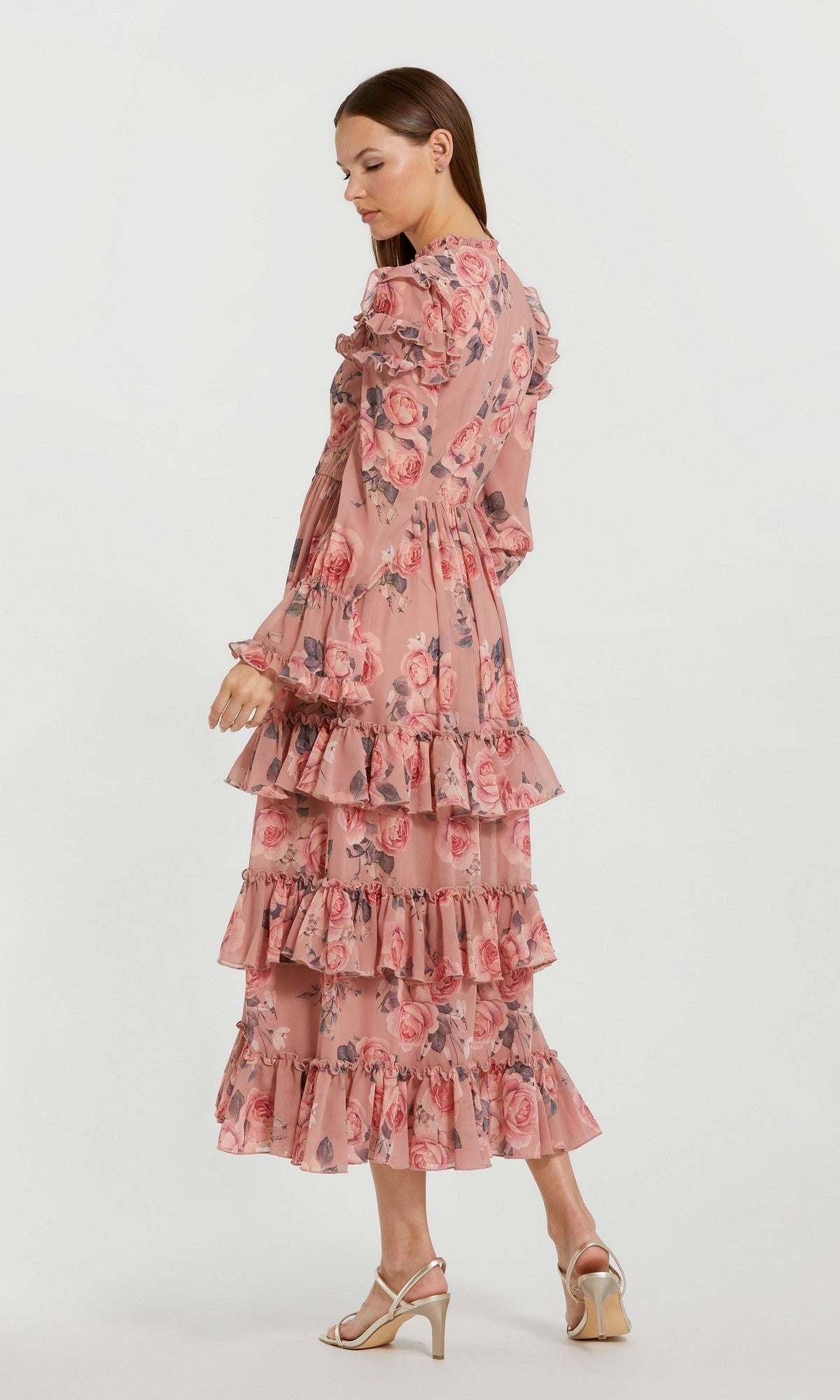 A woman stands sideways against a plain white background wearing the Mac Duggal 9306 Midi Wedding Guest Dress, a long-sleeved, tiered pink floral chiffon dress with ruffles.