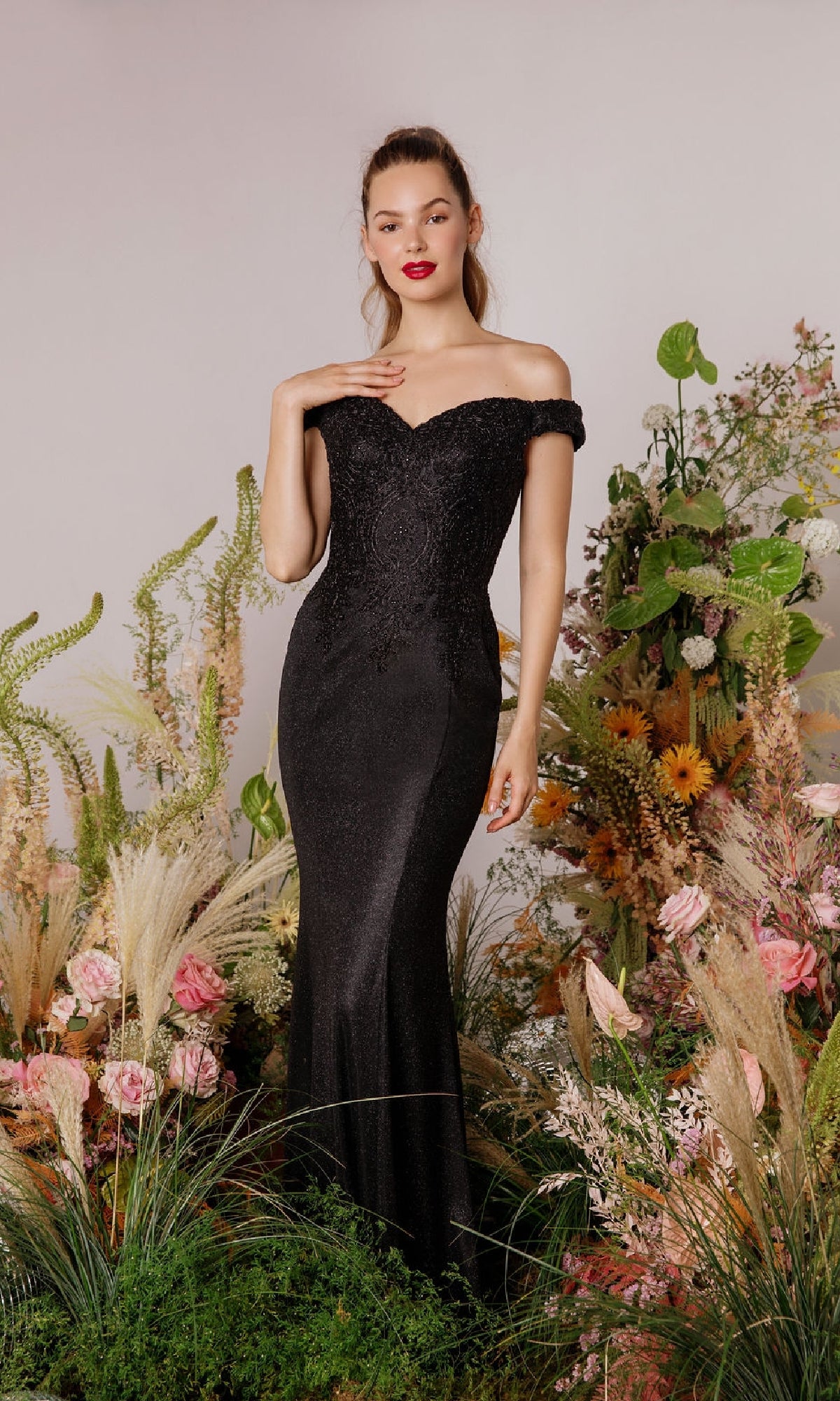 A woman stands among lush greenery and colorful flowers in the Off-Shoulder Long Glitter Prom Dress: Eureka 9306, the fitted black gown making an elegant statement against a plain background.