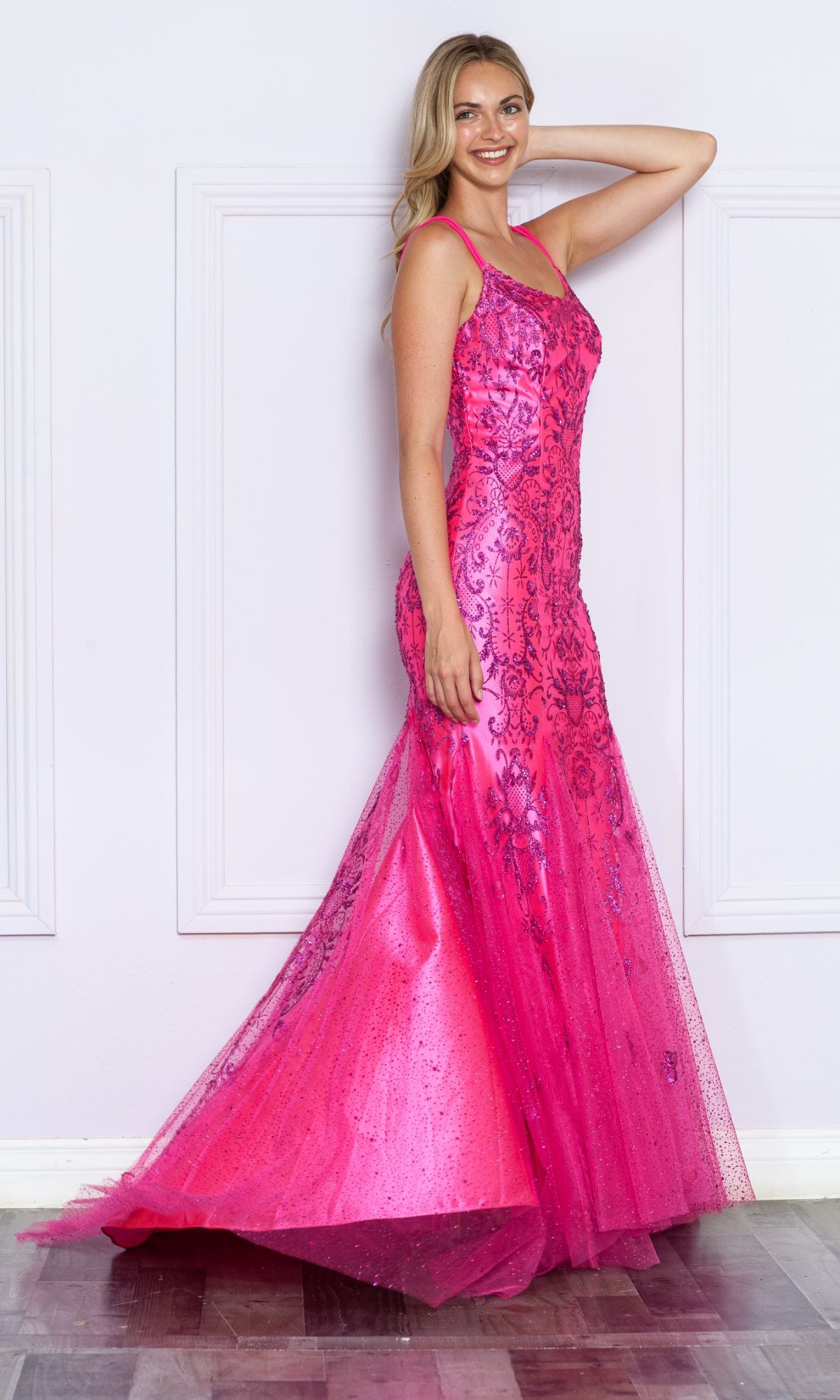 A woman indoors models the Glitter-Print Long Mermaid Prom Dress 9306, featuring a vibrant pink color, sequins, a flared tulle skirt, and thin straps. She smiles with one hand raised, highlighting the gown’s glamorous style.