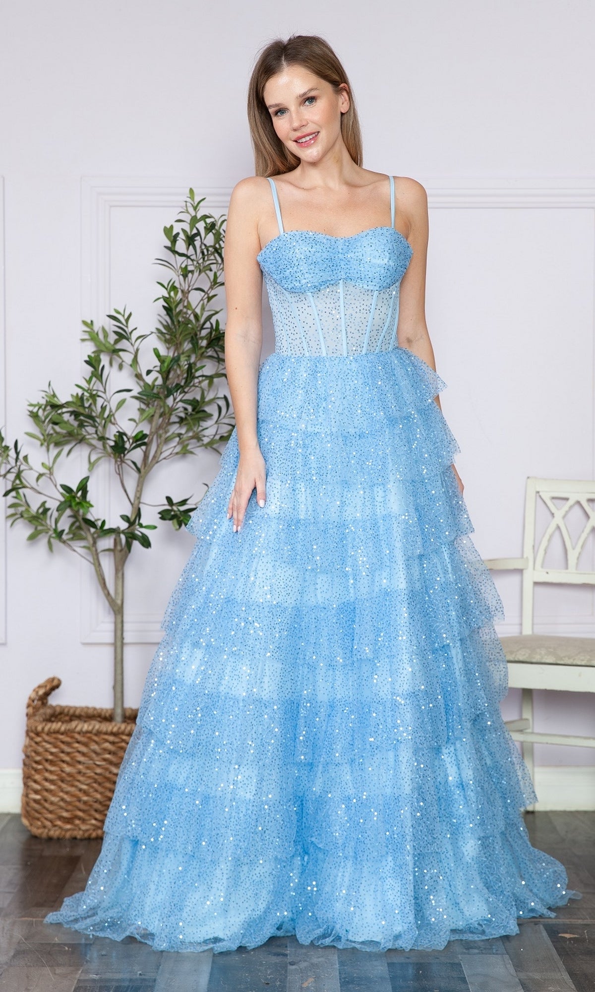 A woman stands indoors by a potted plant and white bench, wearing the Corset-Back Long Tiered Sequin Prom Ball Gown 9328 in sparkling light blue with thin straps and a corset bodice.