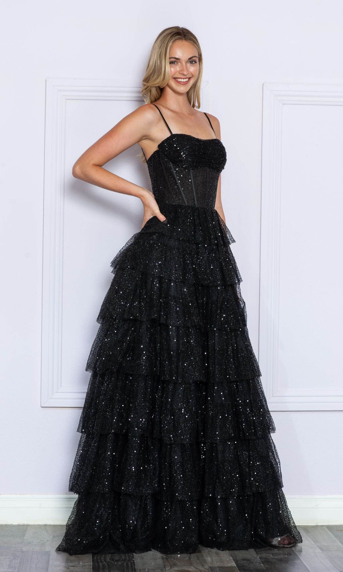 A woman stands indoors wearing the Corset-Back Long Tiered Sequin Prom Ball Gown 9328, a black, floor-length dress with thin straps and a fitted corset bodice.