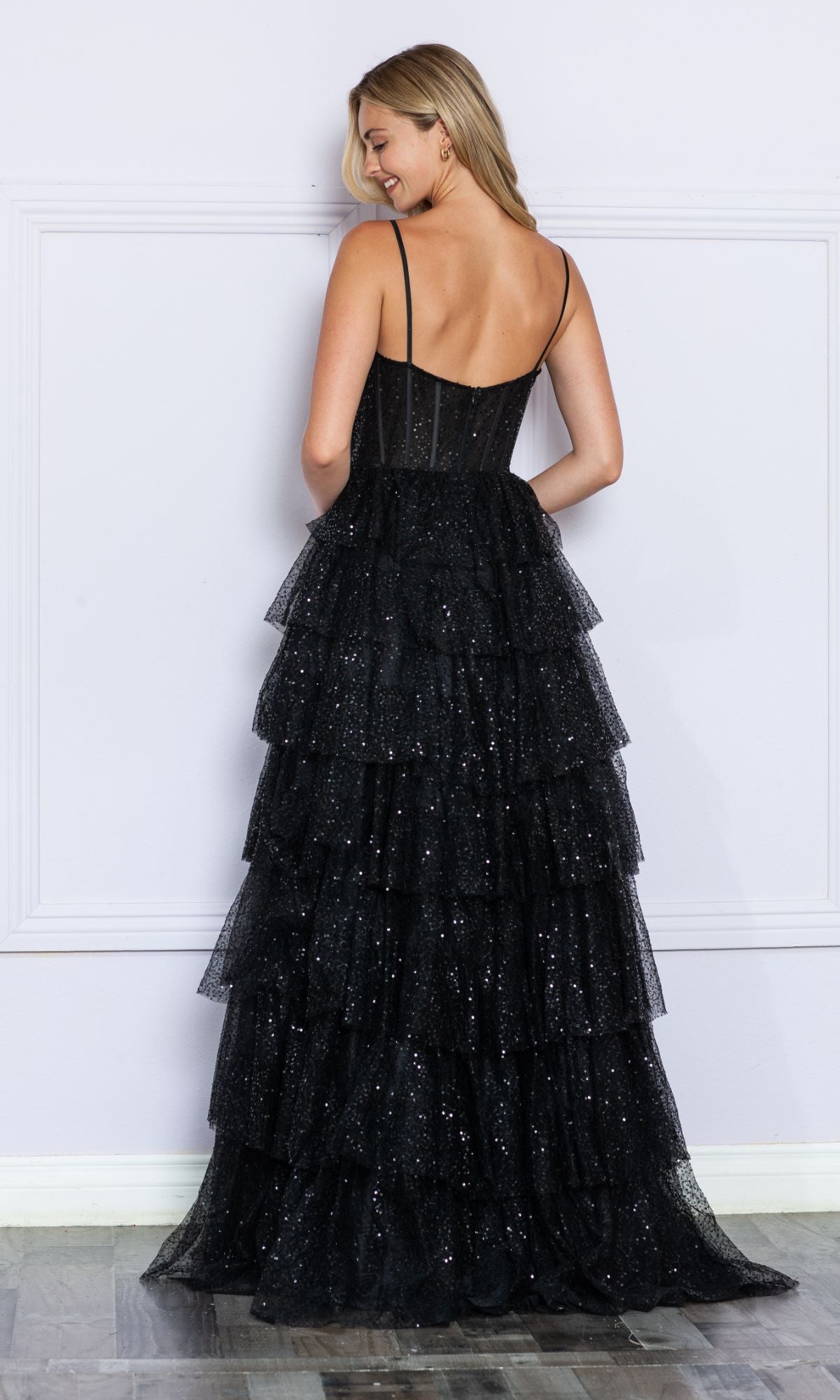 A woman faces away, wearing the Corset-Back Long Tiered Sequin Prom Ball Gown 9328—its black sequin corset bodice and thin straps, plus a sparkling tiered skirt, stand out against a light-colored wall.
