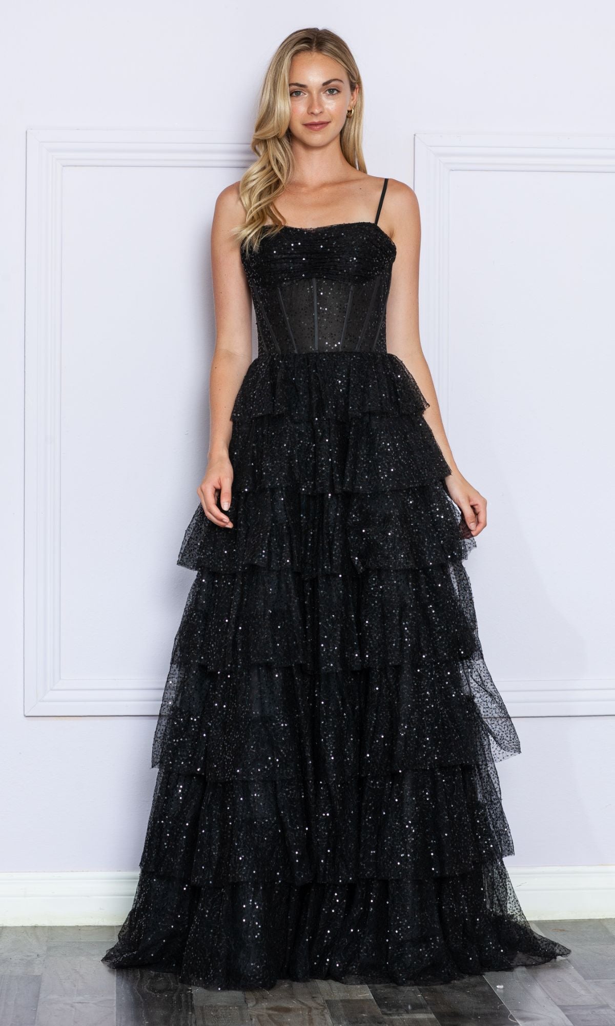 A woman poses against a light wall in the Corset-Back Long Tiered Sequin Prom Ball Gown 9328, a sleeveless, floor-length black dress with tiered ruffles and glittering sequin details.