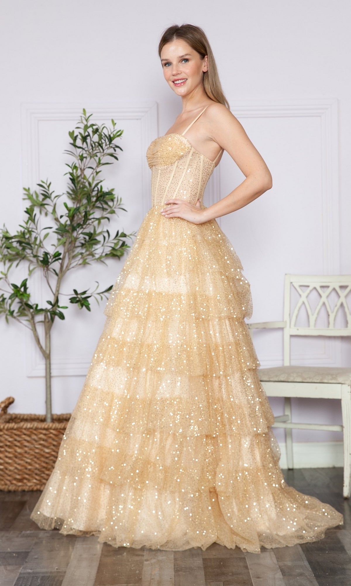 A woman poses indoors in front of a white wall, potted plant, and chair, wearing the Corset-Back Long Tiered Sequin Prom Ball Gown 9328—a sparkling gold dress with spaghetti straps and a corset bodice.