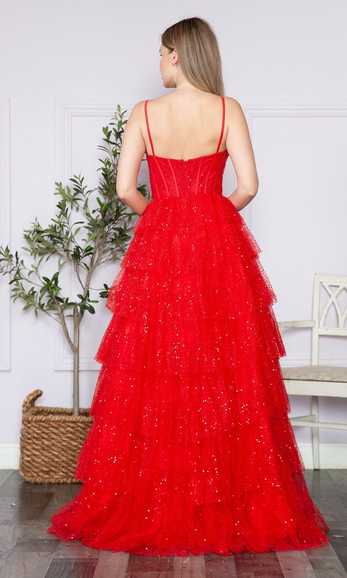 A woman stands indoors, shown from the back, wearing the Corset-Back Long Tiered Sequin Prom Ball Gown 9328 with thin straps and a corset bodice. A plant and a white bench are in the background.