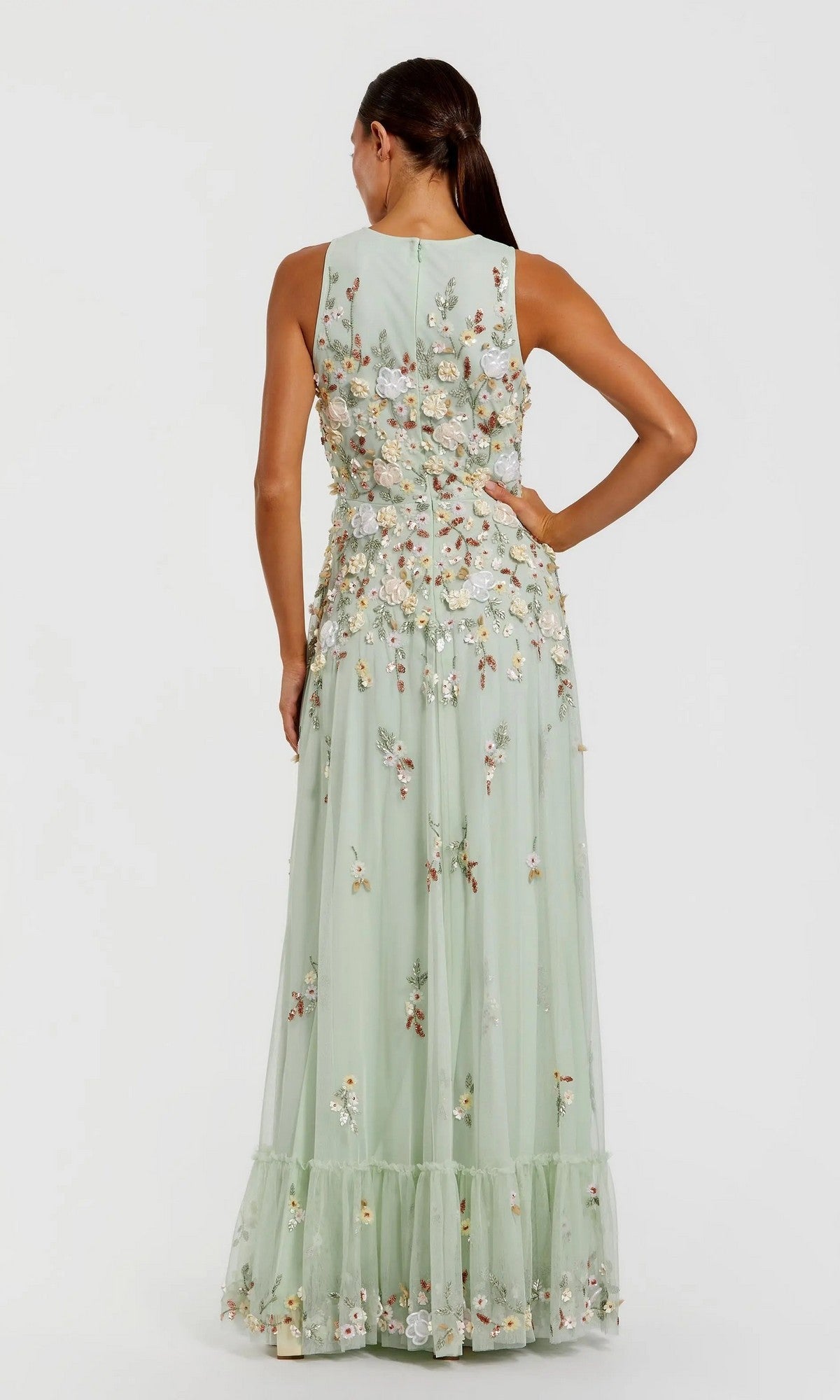 Against a plain background, a woman stands with her back to the camera wearing the Mac Duggal 9334 Long Formal Dress—a sleeveless, light green gown adorned with floral embroidery and a sheer overlay.