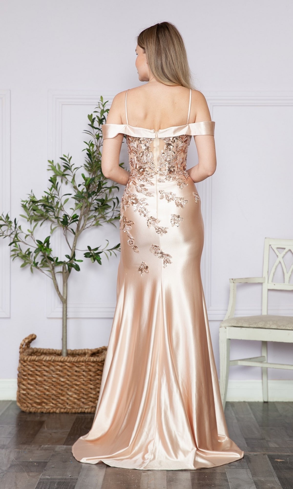 A woman faces away from the camera indoors in the Embellished Off-the-Shoulder Long Prom Dress 9350, a champagne-colored gown with floral embroidery, shimmering near a potted plant and white chair.