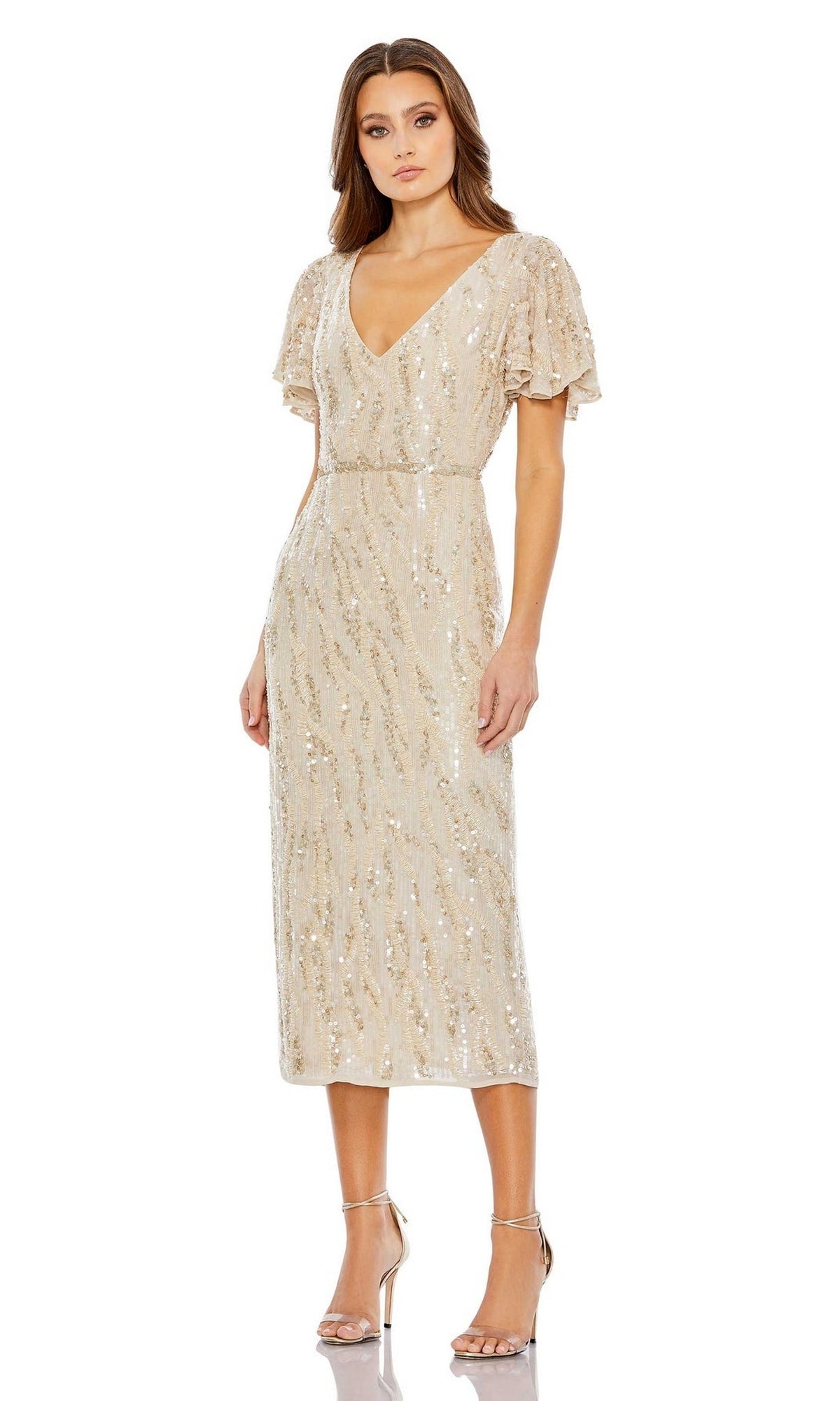 A woman models the Mac Duggal Short Wedding Guest Dress 93591, a light beige fitted party dress with sequin embellishments, V-neckline, and short flutter sleeves, paired with silver high-heeled sandals against a white background.