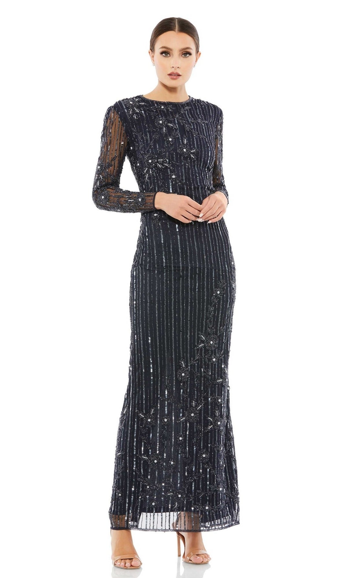 A woman stands wearing the Mac Duggal 93626 Long Sleeve Sequin Formal Gown—a floor-length black dress with silver sequin details and sheer fabric—paired with nude high-heeled sandals.