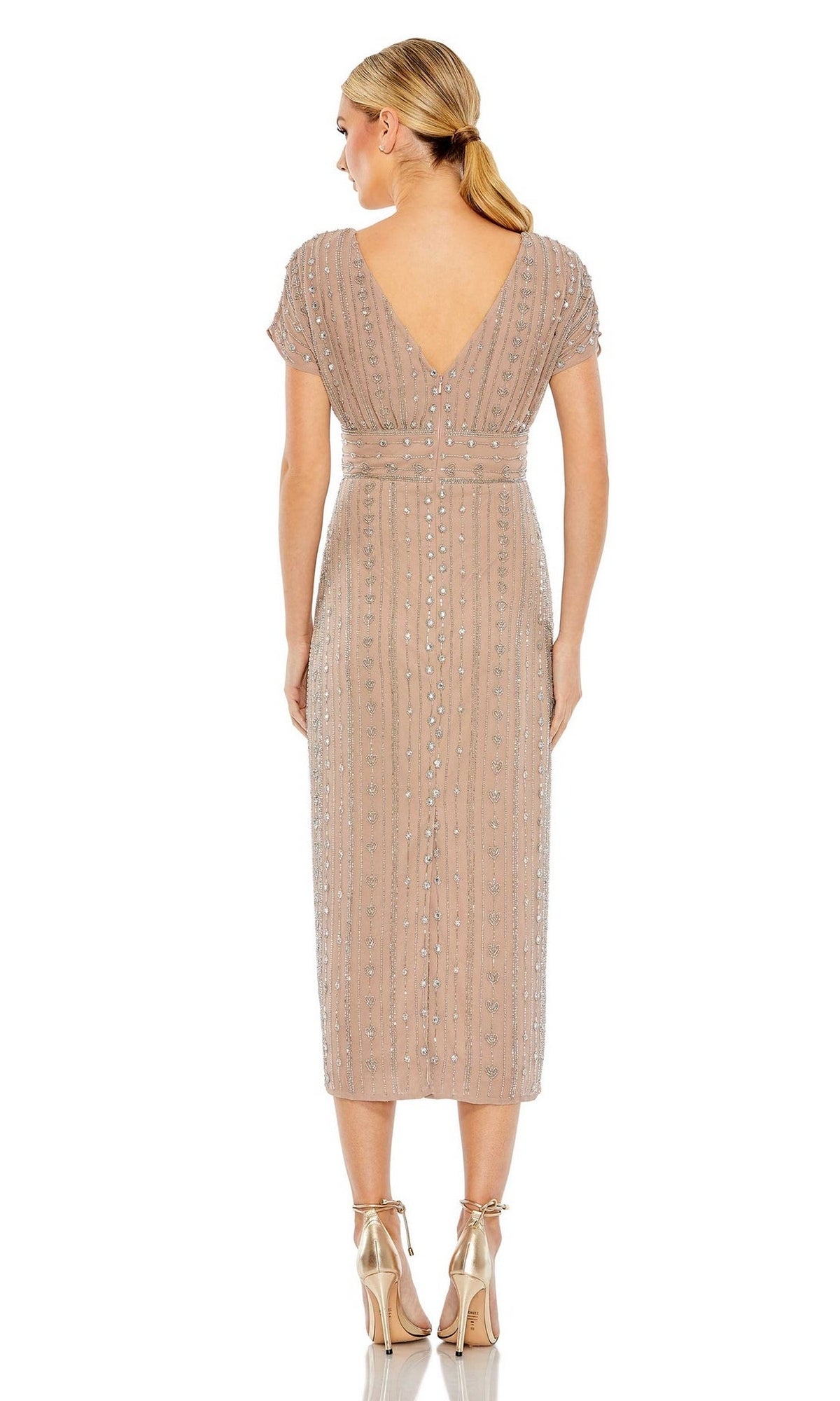 A woman faces away, wearing the Mac Duggal Beaded Short Wedding-Guest Dress 93649 in light beige with short sleeves and metallic high heels. Her hair is styled in a low ponytail.