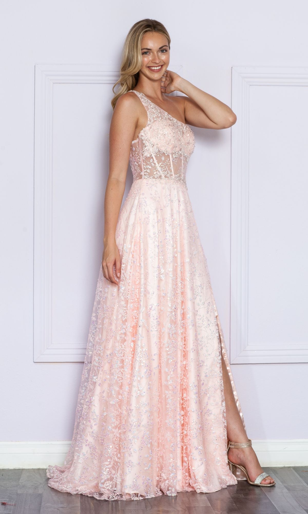 A woman poses indoors in the One-Shoulder Embroidered Long Prom Dress 9372—a light pink formal gown with a slit, sparkling embellishments, and elegant embroidery—wearing one hand on her head and the other by her side.