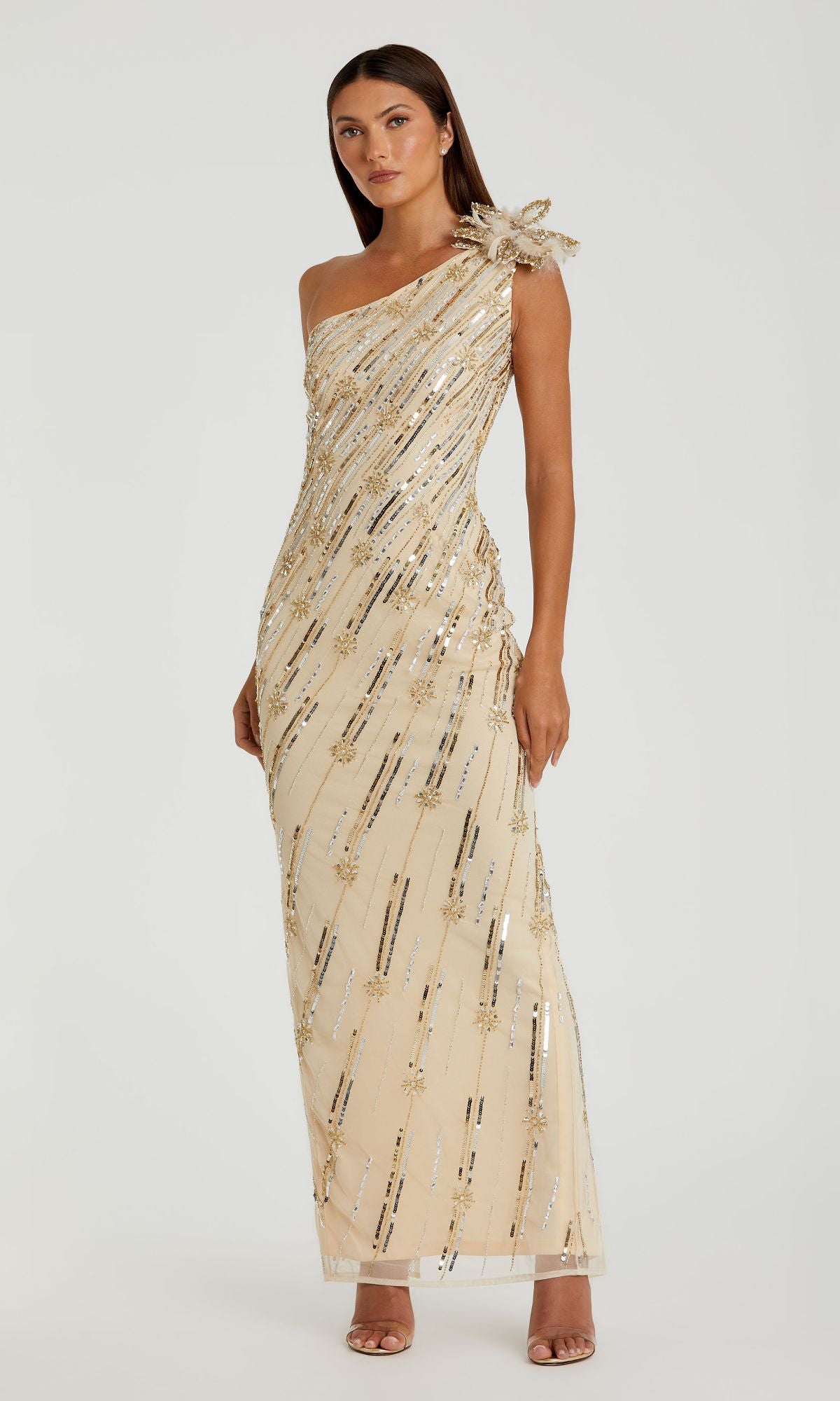 A woman models the Mac Duggal 93739 Long Gold Sequin Formal Dress, featuring a one-shoulder design with sequin detailing and a bow, paired with strappy heels against a plain background.