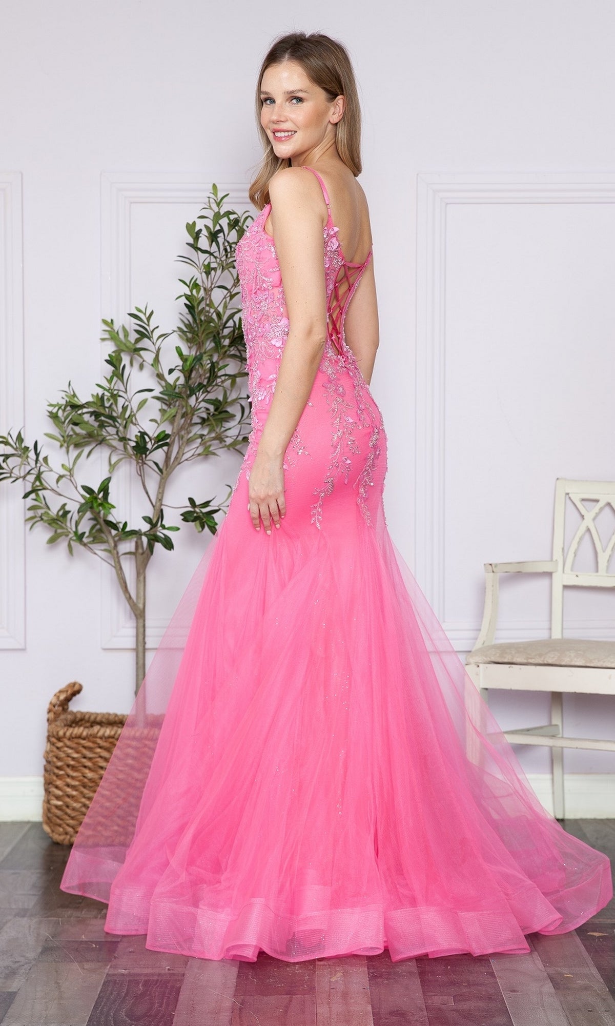 A woman stands indoors, smiling over her shoulder in the Sheer-Corset-Bodice Long Mermaid Prom Dress 9374—a floor-length pink gown with lace-up back and delicate floral embroidery.