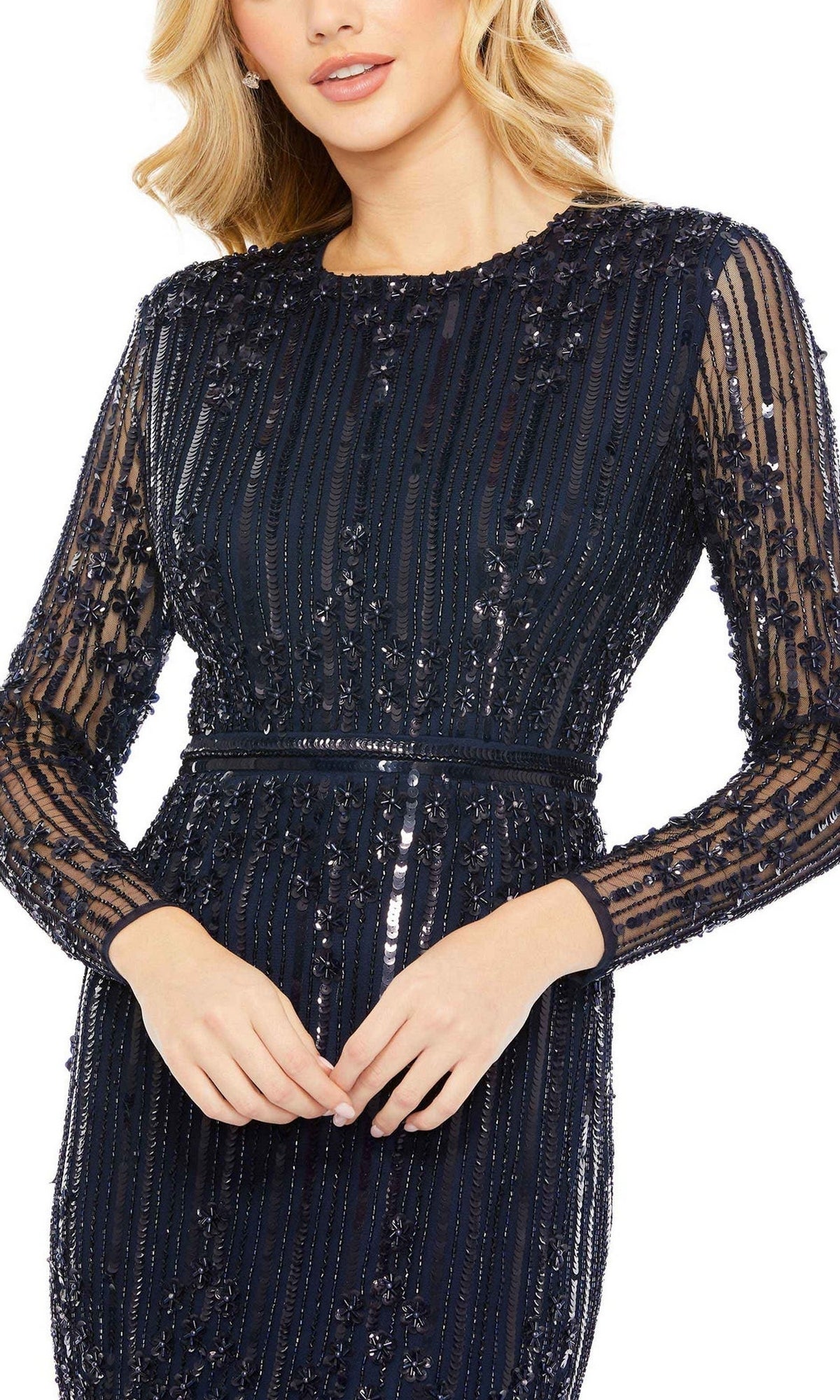 A woman poses with hands clasped, wearing the Mac Duggal 93781 Long Sleeve Beaded Formal Gown in navy, featuring sheer long sleeves and intricate bead detailing.