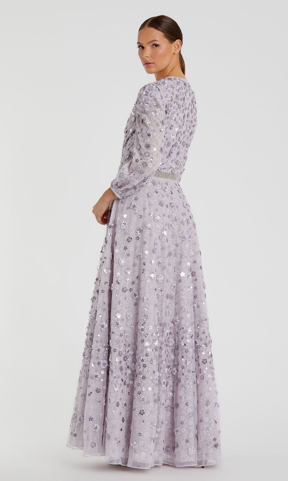 A woman poses in the Mac Duggal 93805, a modest long sleeve formal dress in lavender with sequin embellishments, looking over her shoulder against a plain white background.