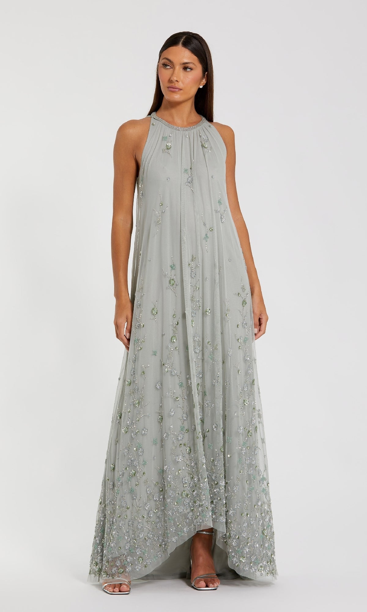 A woman stands against a white background, wearing the Mac Duggal 9382— a sage green, floor-length formal dress with delicate floral embroidery and no sleeves.
