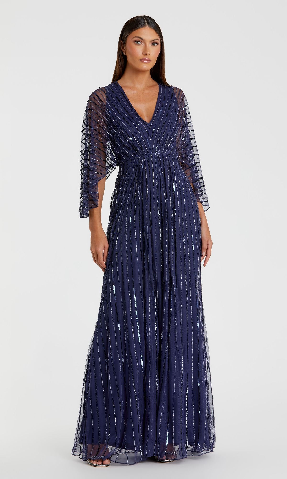 A woman stands against a plain white background, wearing the Mac Duggal 93841—a Long Sequin A-Line Formal Dress featuring navy blue sequins, sheer sleeves, and vertical stripes.