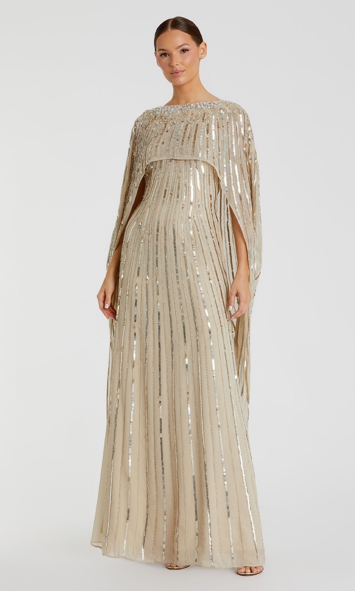 A woman wears the Caped Long Sequin Formal Dress (Mac Duggal 93869), a floor-length beige gown with vertical silver sequin stripes and a cape overlay, standing against a plain white background.