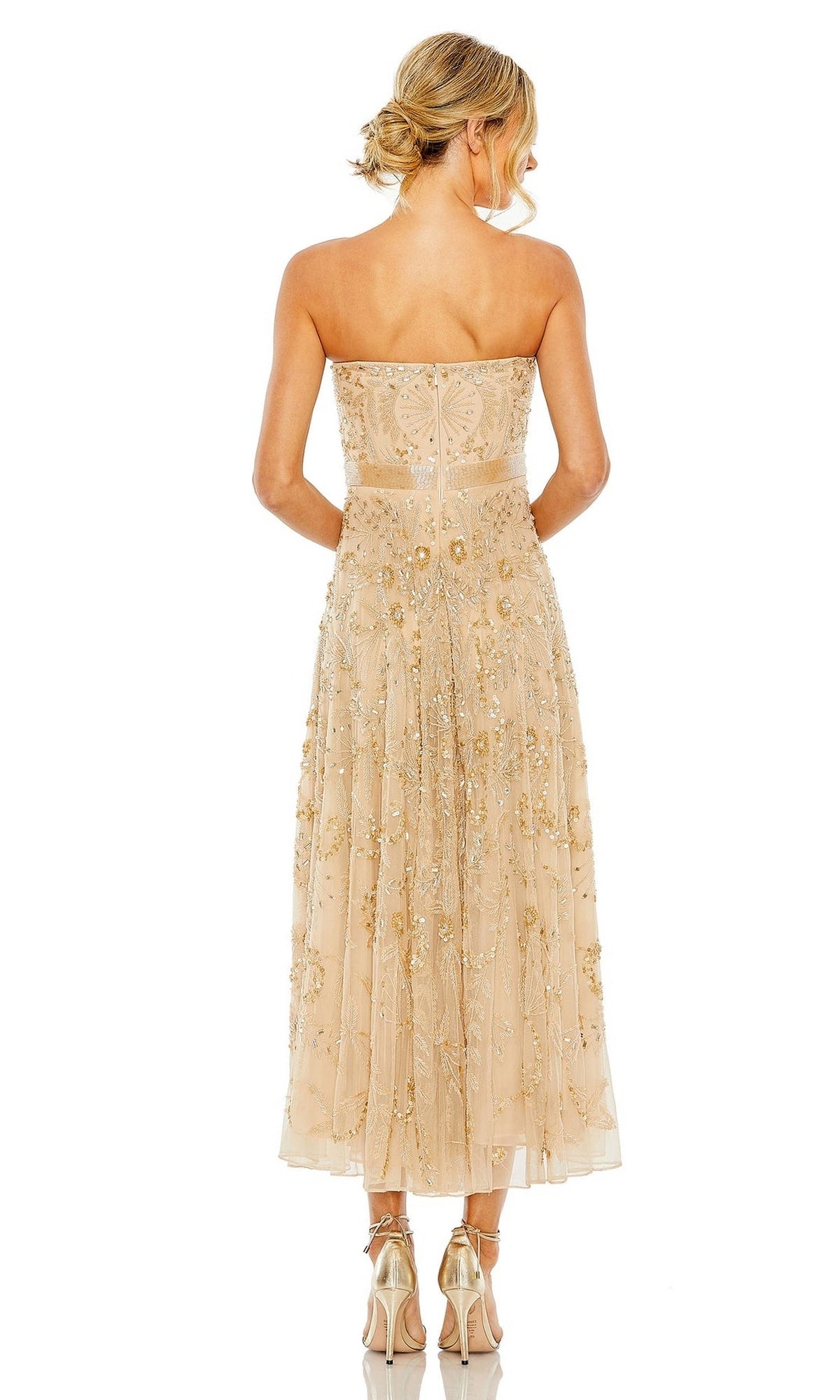 A woman facing away wears the Mac Duggal 93898 Strapless Wedding Guest Dress with a beaded design, sheer overlay, and metallic heels. Her blonde hair is styled in a loose updo.