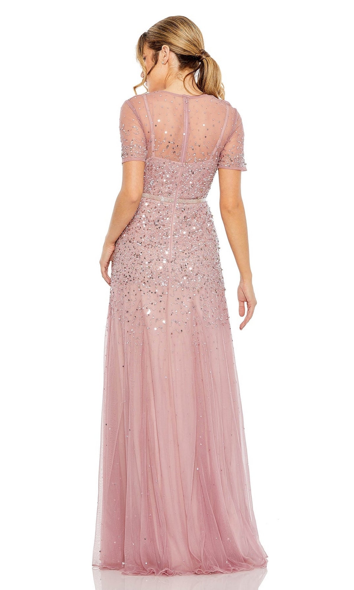 A woman faces away in the Mac Duggal Sheer High-Neck Sequin Formal Dress 93909, a light pink A-line gown with sheer fabric, sparkling embellishments, a beaded waist, and her hair in a low ponytail.