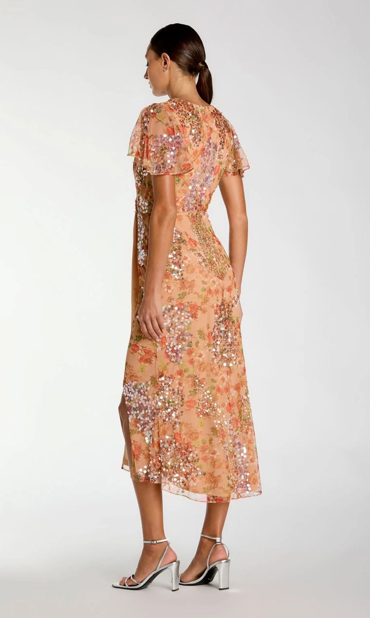 Woman in profile wearing the Mac Duggal 93949 Midi Wedding Guest Dress in sequin peach floral with flutter sleeves, paired with silver block-heeled sandals.