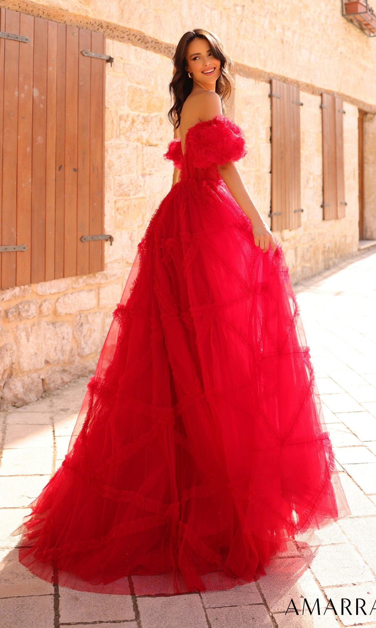 A woman in the Amarra Puff-Sleeve Long Ruffled Prom Dress 94002 stands outdoors on a sunlit stone pathway, smiling over her shoulder in the bright red, off-the-shoulder, floor-length gown.