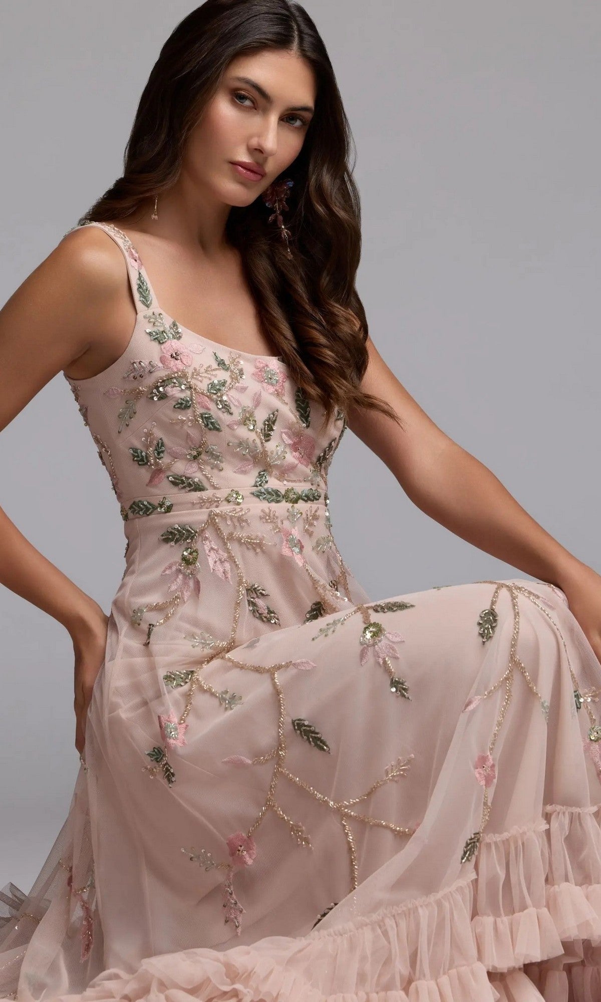 A woman wearing the Mac Duggal 9402 long formal dress, a sleeveless light pink party gown with floral beaded embroidery, is seated against a plain gray background.