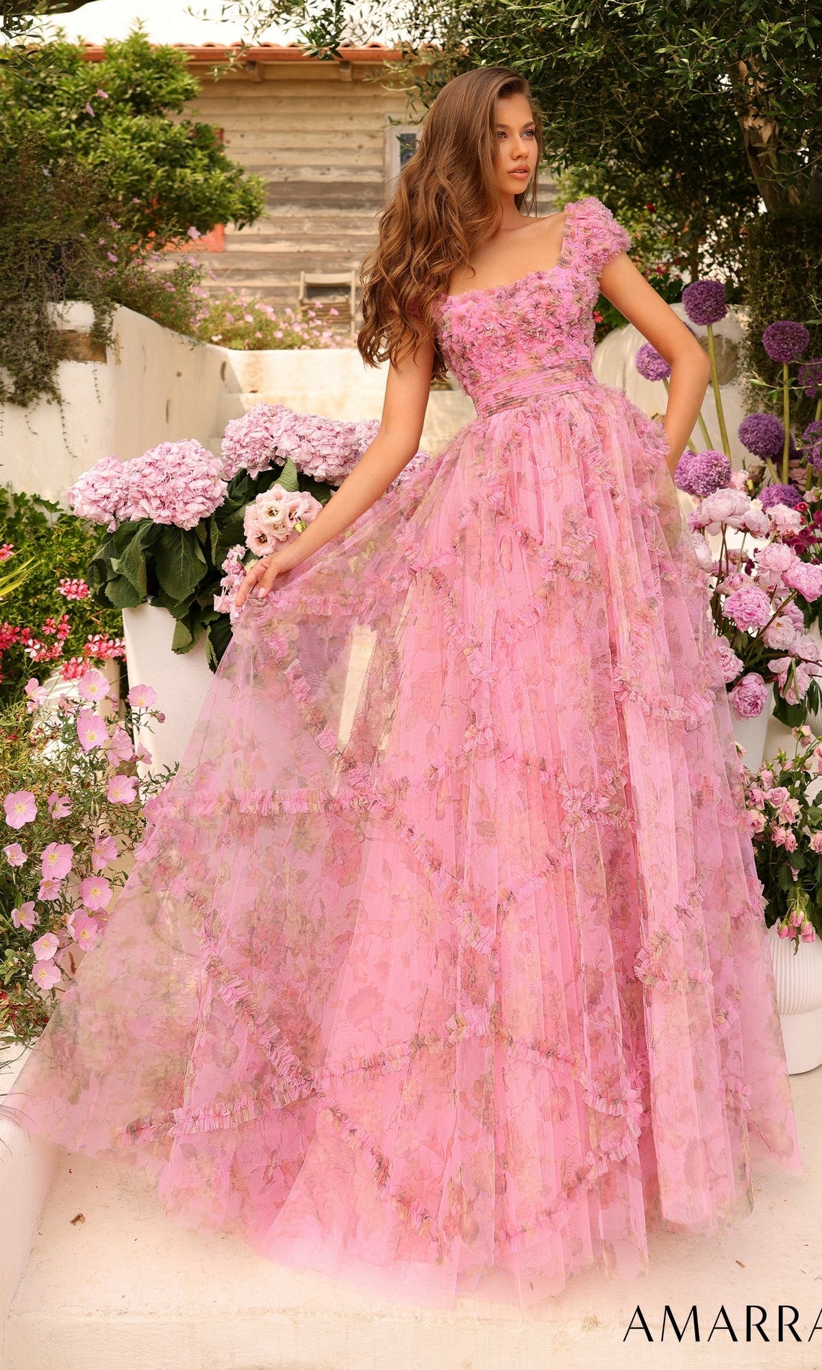 A woman stands in a garden of blooming hydrangeas, wearing the Amarra 94044 Cap-Sleeve Long Ruffled Prom Gown, a floor-length pink floral-print dress with sheer layers and a fitted bodice.