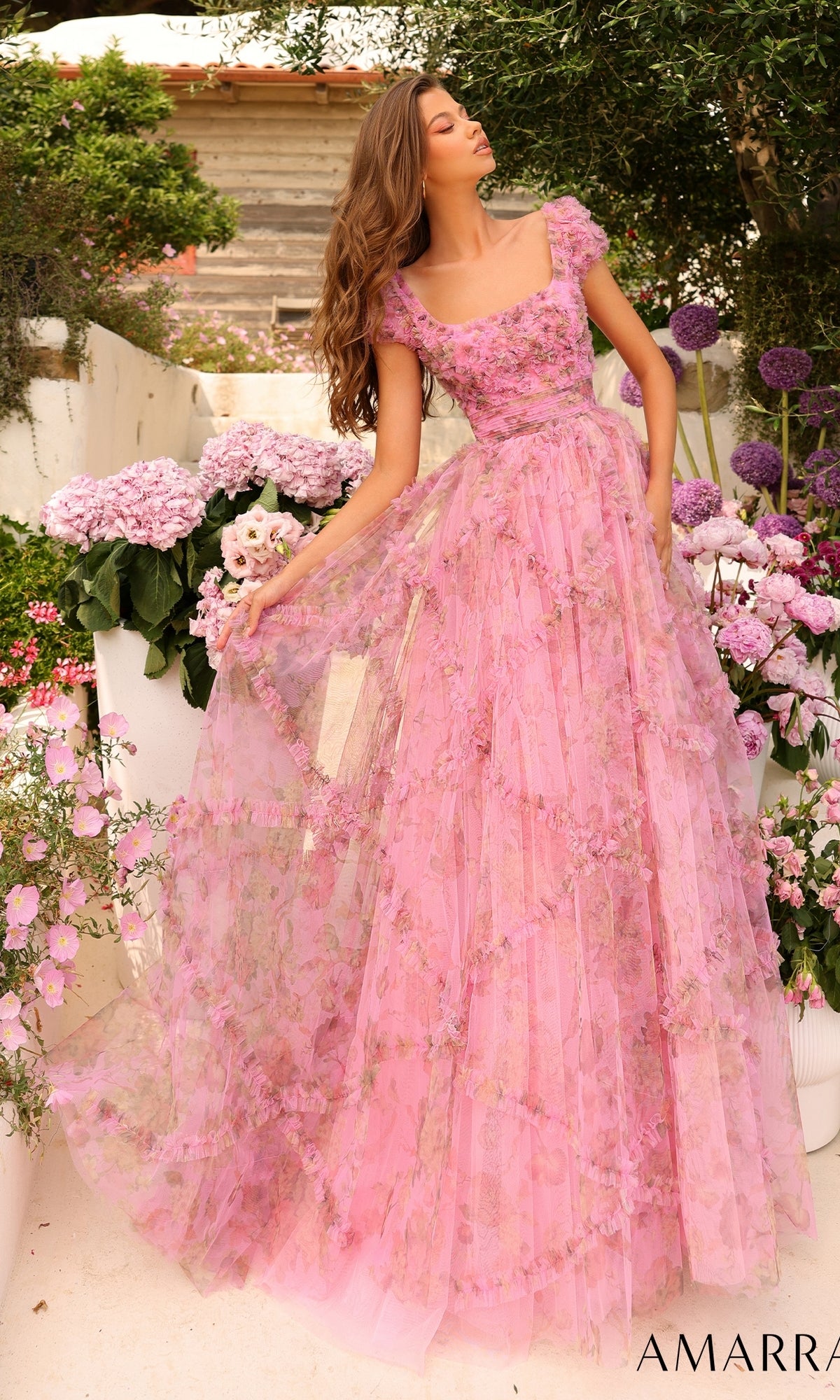 A woman stands outdoors among blooming purple and pink flowers, gracefully holding the skirt of her Cap-Sleeve Long Ruffled Prom Gown: Amarra 94044.