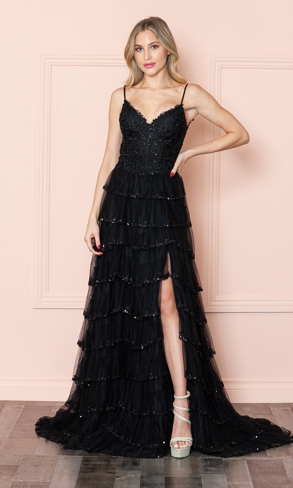 A woman with light hair in strappy heels stands by a pale pink wall, wearing the Ruffled Long A-Line Tiered Prom Dress: Poly 9404—a black evening gown with thin straps, sequin accents, ruffles, and a slit.