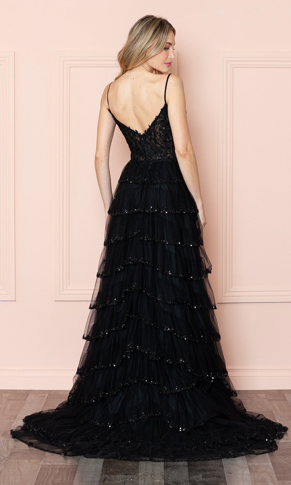 A woman in the Ruffled Long A-Line Tiered Prom Dress: Poly 9404, a black floor-length gown with spaghetti straps and a deep V-back, stands facing away against a pale pink wall.