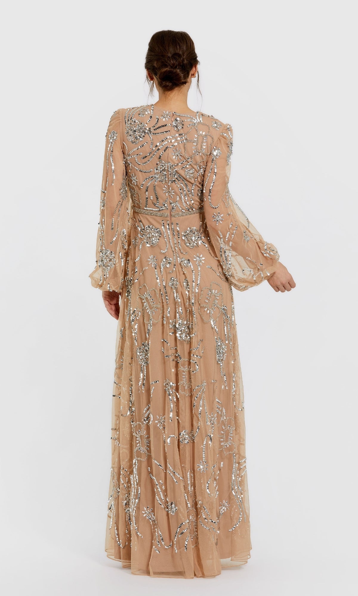 A woman with brown hair in an updo models the Mac Duggal 9472, a long sleeve nude formal gown adorned with silver sequins, shown from the back against a plain light background.