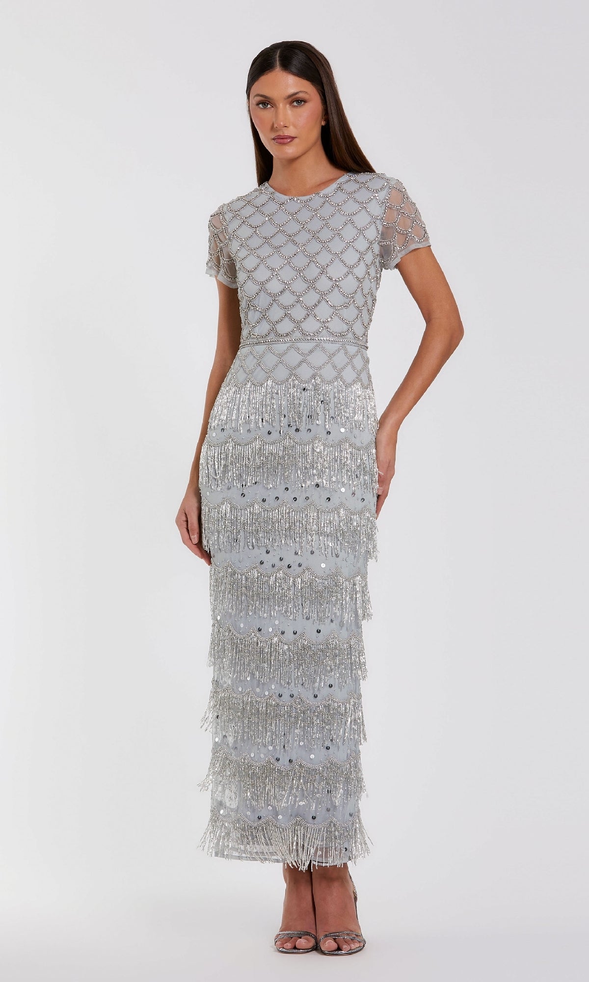A woman stands against a plain white background wearing the Mac Duggal 94077 Fringed Short Sleeve Party Dress—a silver, floor-length gown with beaded geometric patterns and tiered fringe.