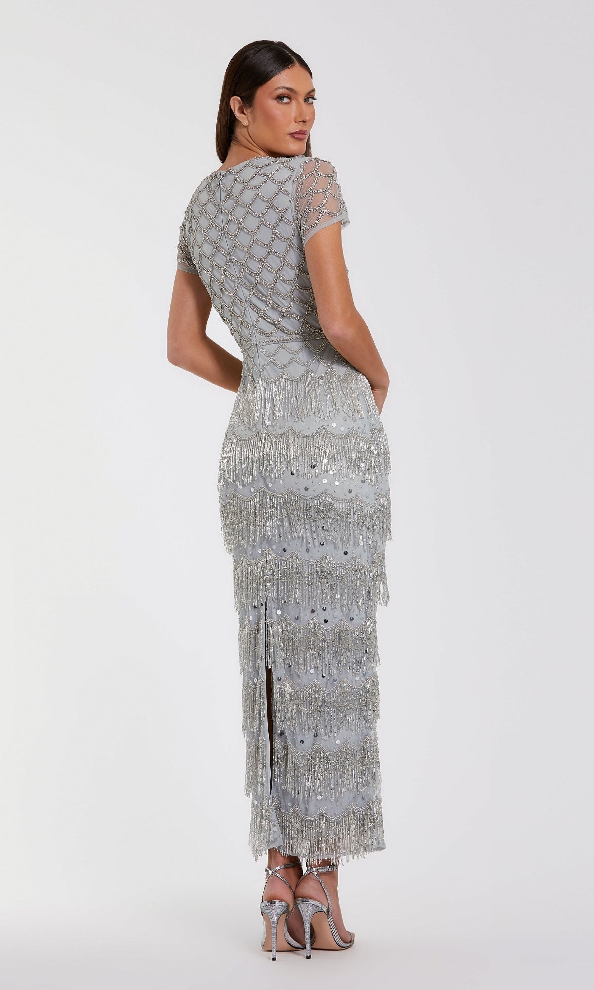 A woman stands with her back to the camera in the Mac Duggal 94077 Fringed Short Sleeve Party Dress, a silver floor-length gown shimmering with beading, intricate embellishments, and long dramatic fringe.
