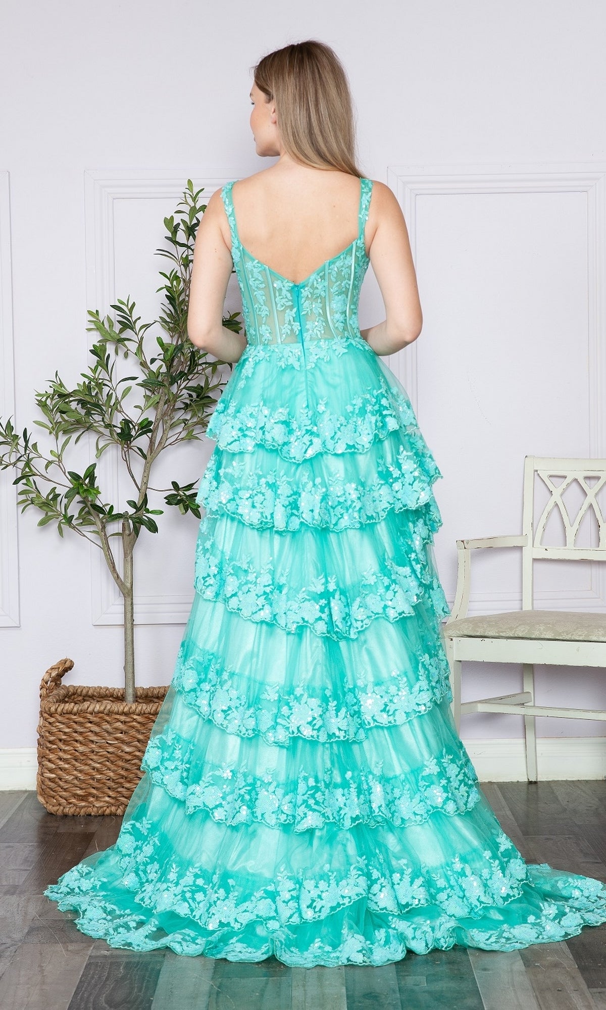 A woman in the Sheer-Corset Long Ruffled Prom Dress (Poly USA 9410), a turquoise tiered lace gown with a corset bodice, stands with her back to the camera in a bright room featuring light walls, a white chair, and a potted plant.