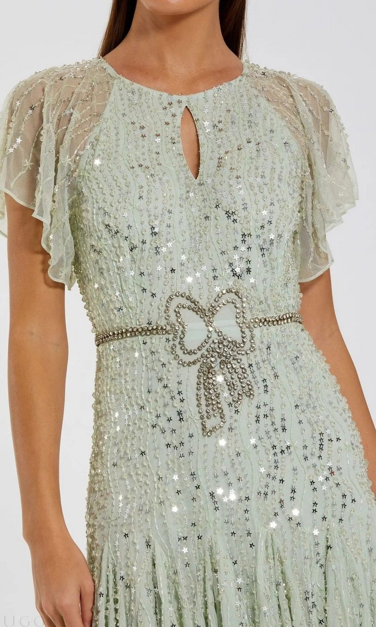 A woman wearing a light green, star sequin tea-length party dress featuring sheer flutter sleeves and a beaded bow-shaped belt, reminiscent of the elegant Mac Duggal 94128.