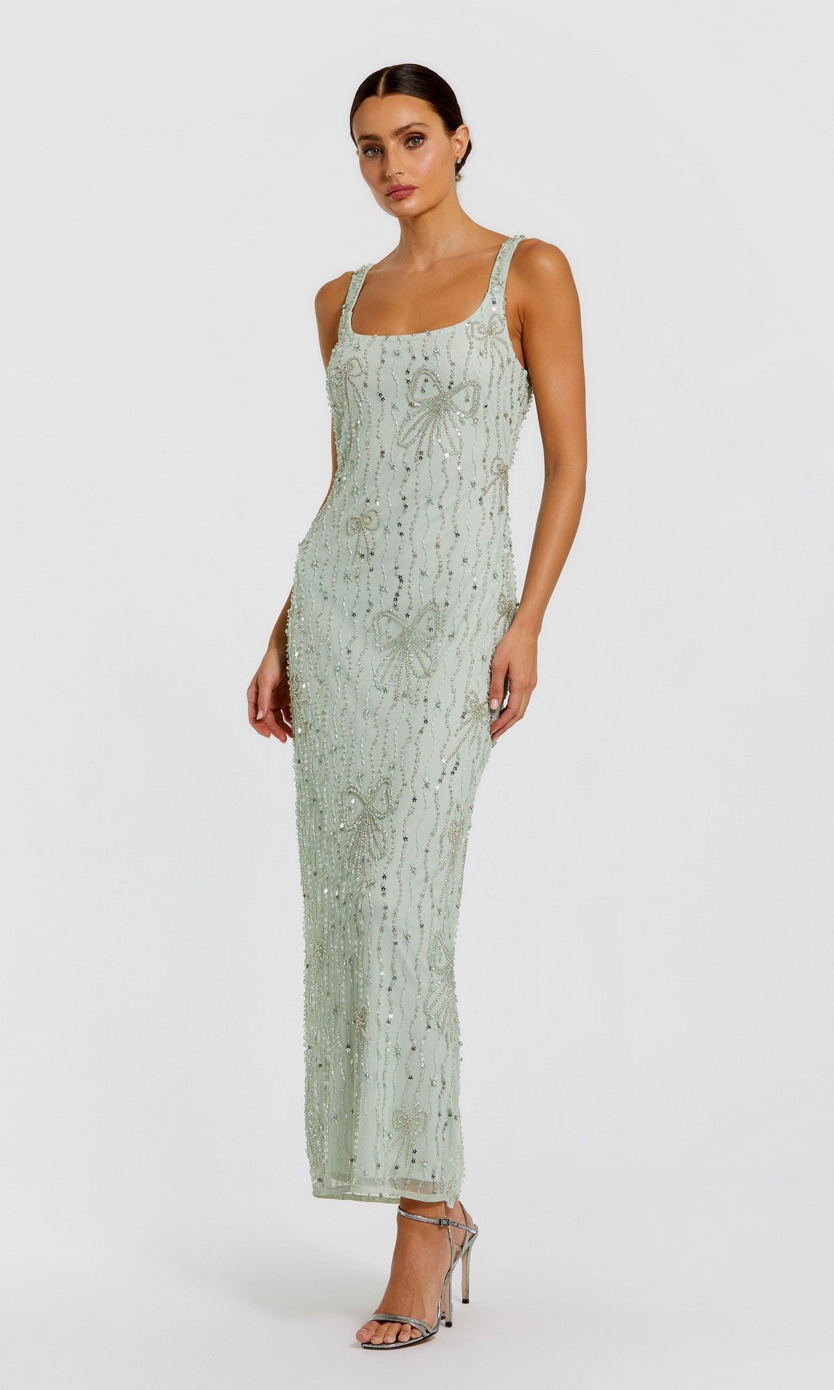 A woman in the Mac Duggal 94129 long formal dress in seafoam, featuring a beaded mesh design with sequin embellishments, and silver high-heeled sandals stands against a plain white background.