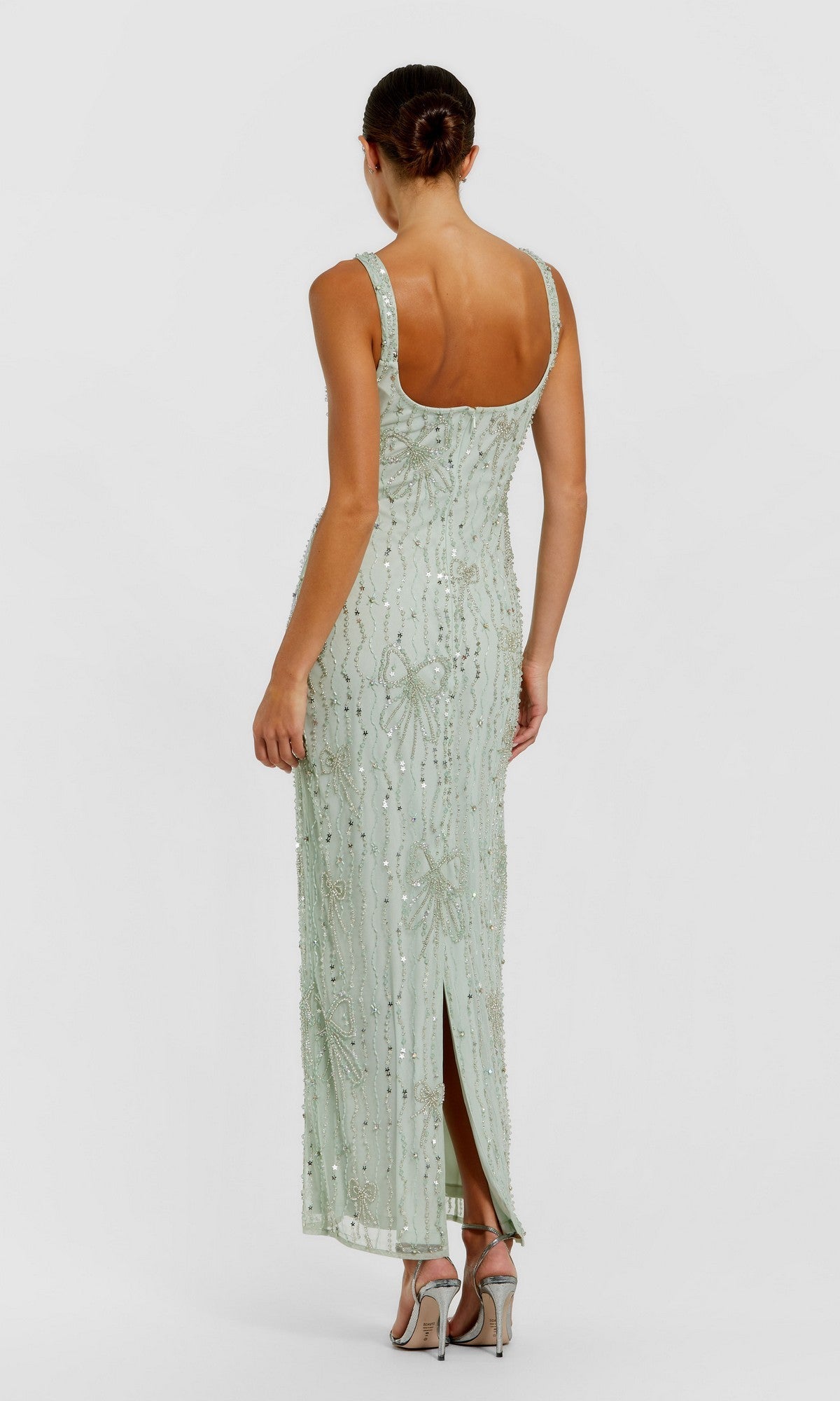 A woman faces away from the camera wearing the Mac Duggal 94129, a pale green sleeveless long formal dress with beaded mesh, subtle sequin accents, and a back slit.